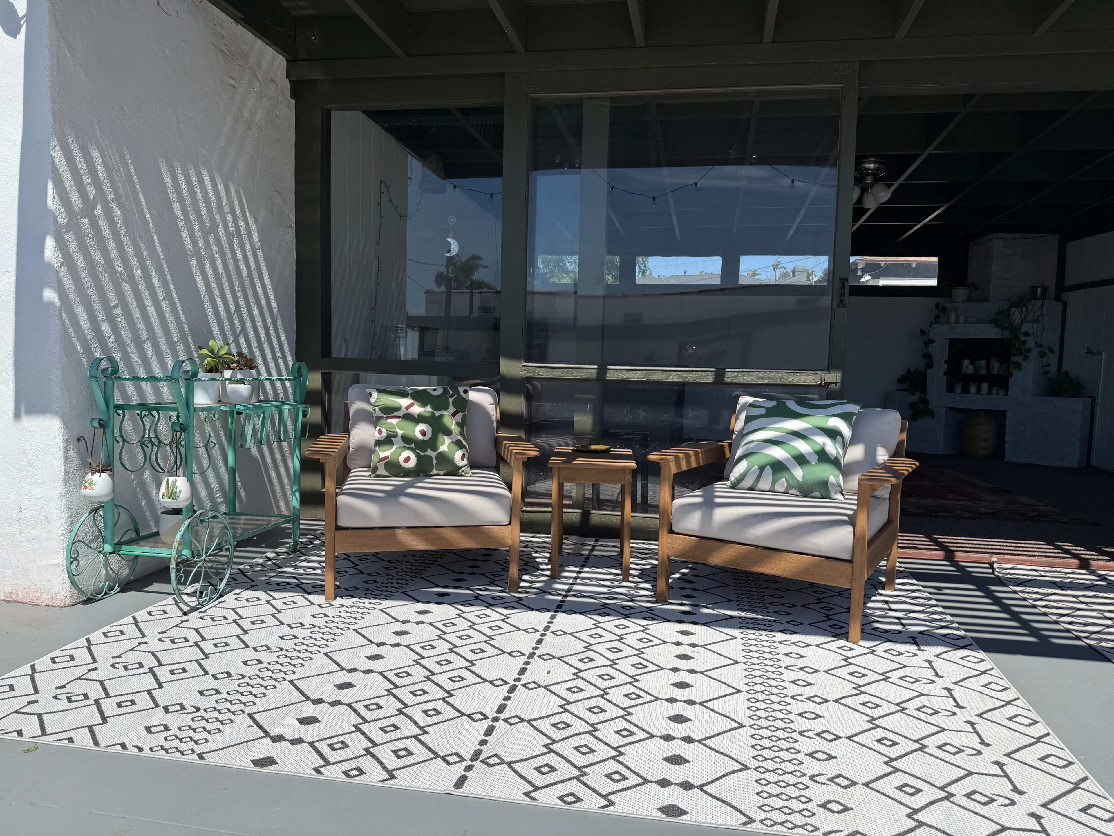 Two wooden chairs with cushions are placed on a patterned rug in a shaded outdoor space.