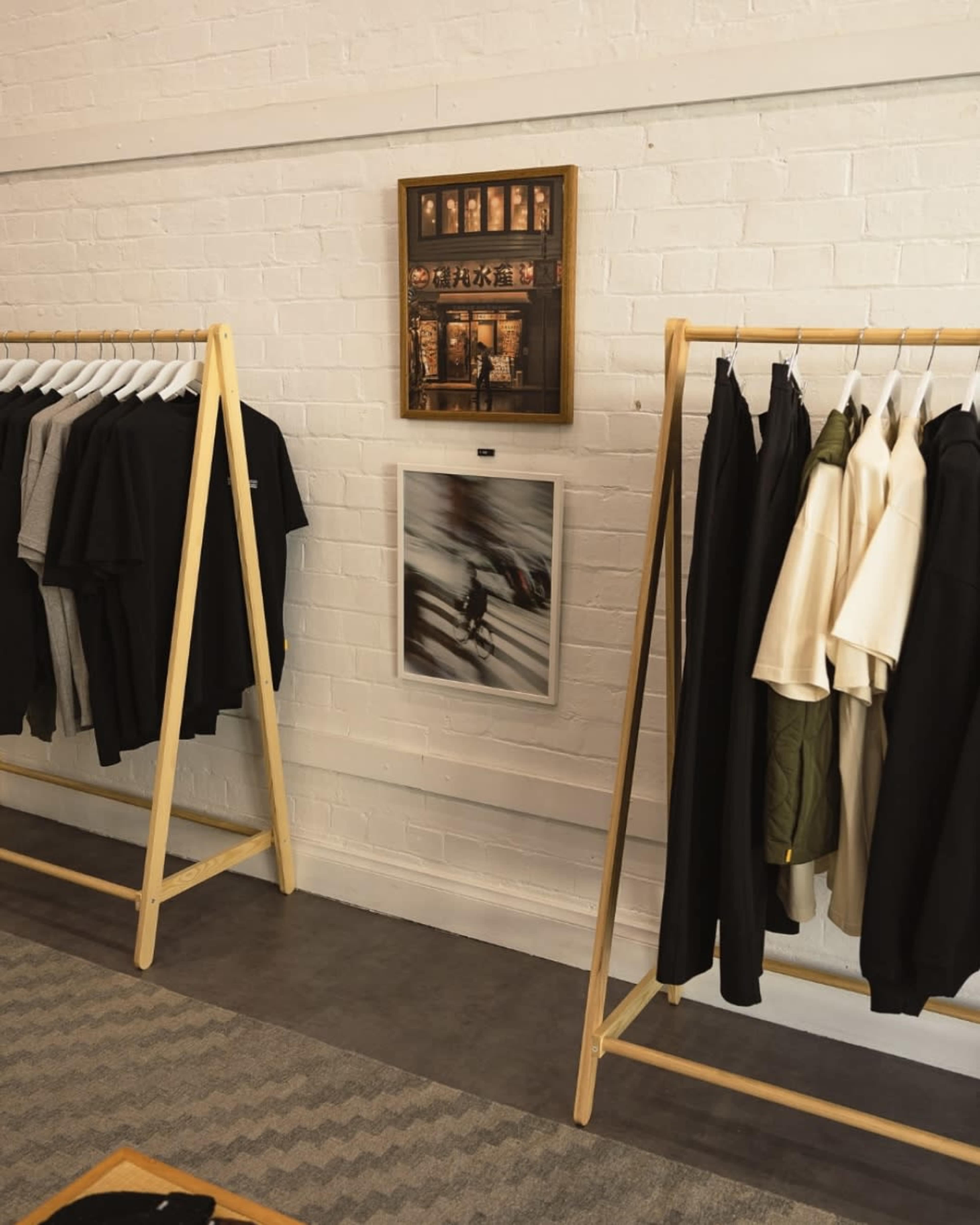 The image shows a clothing retail space featuring wooden racks with various shirts displayed and framed artwork on the wall.