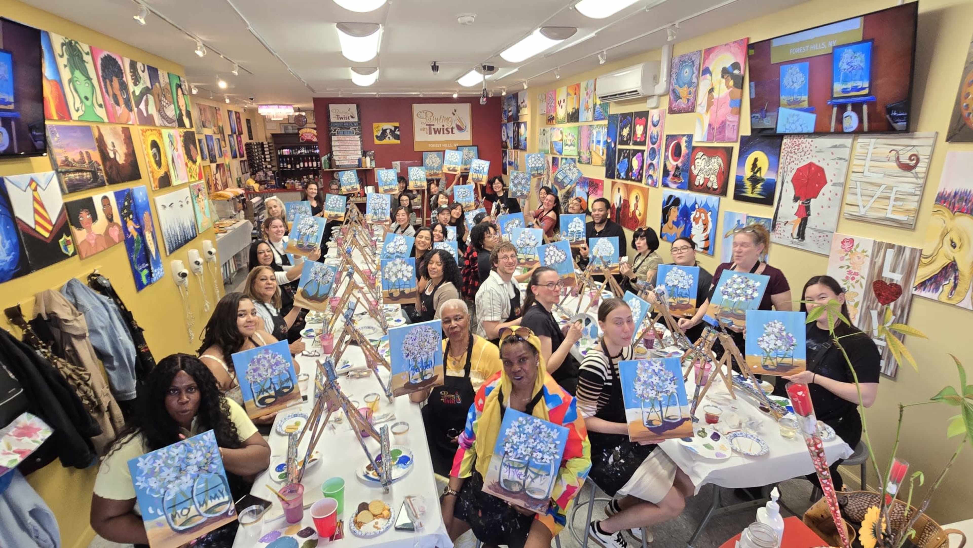A large group of people holds up painted canvases in a brightly decorated art studio filled with various artworks on the walls.