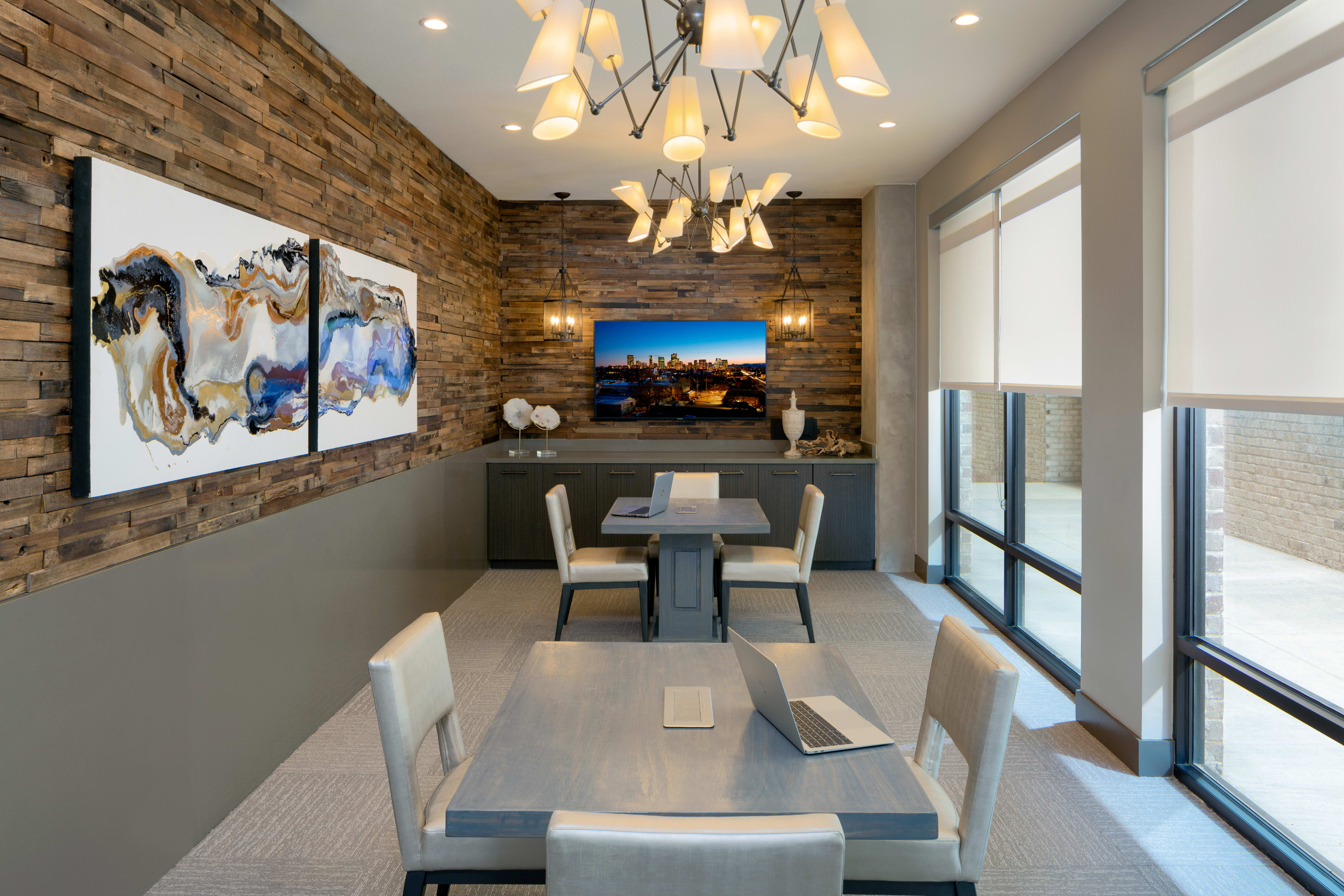 A modern conference room features a wooden accent wall, contemporary lighting, two artworks, a television, and four tables with chairs.