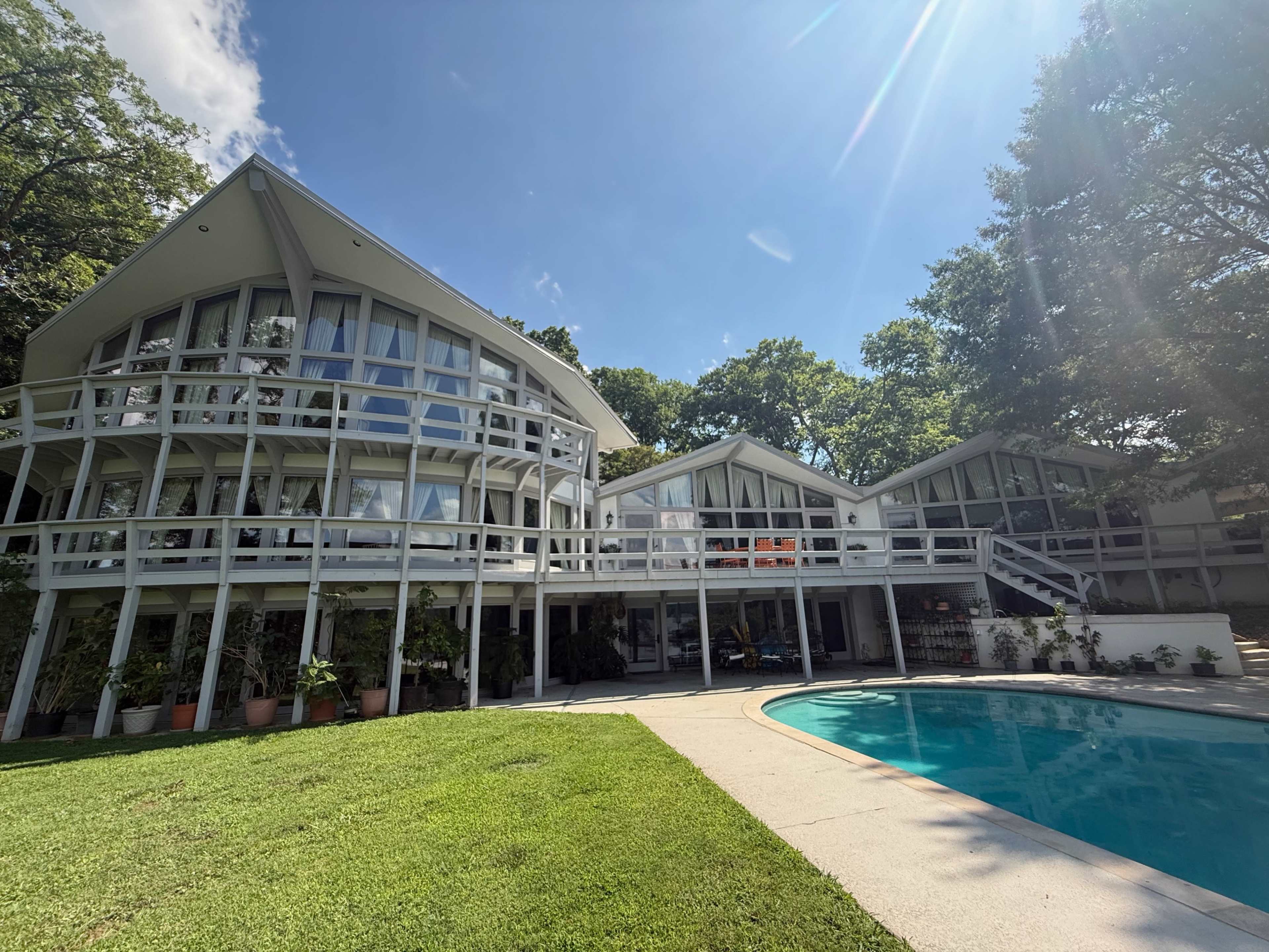 A modern two-story house with large windows and a curved design is situated next to a swimming pool surrounded by a grassy area and tall trees.