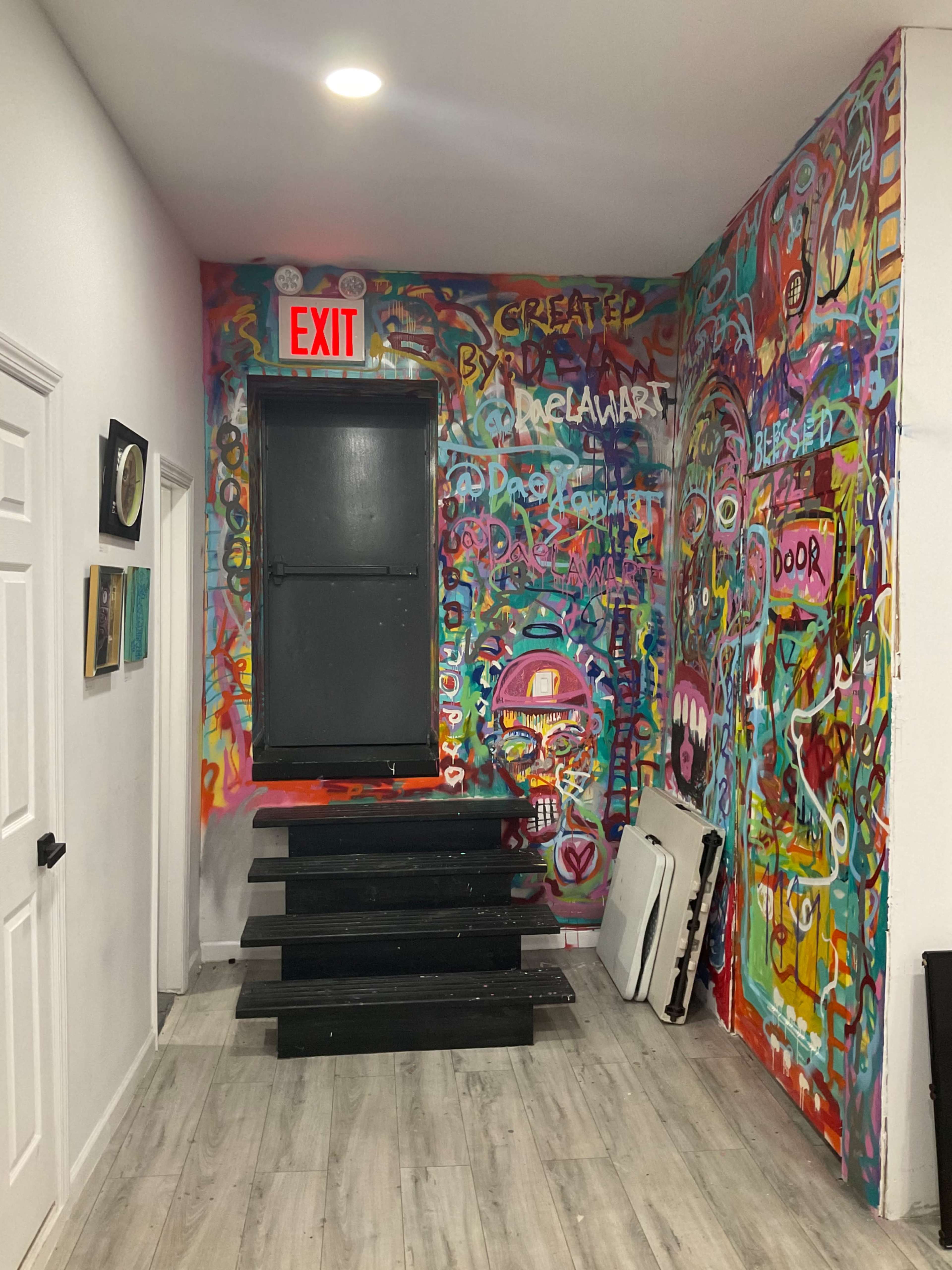 A brightly colored hallway with graffiti-covered walls and a staircase leading to a door marked with an exit sign.