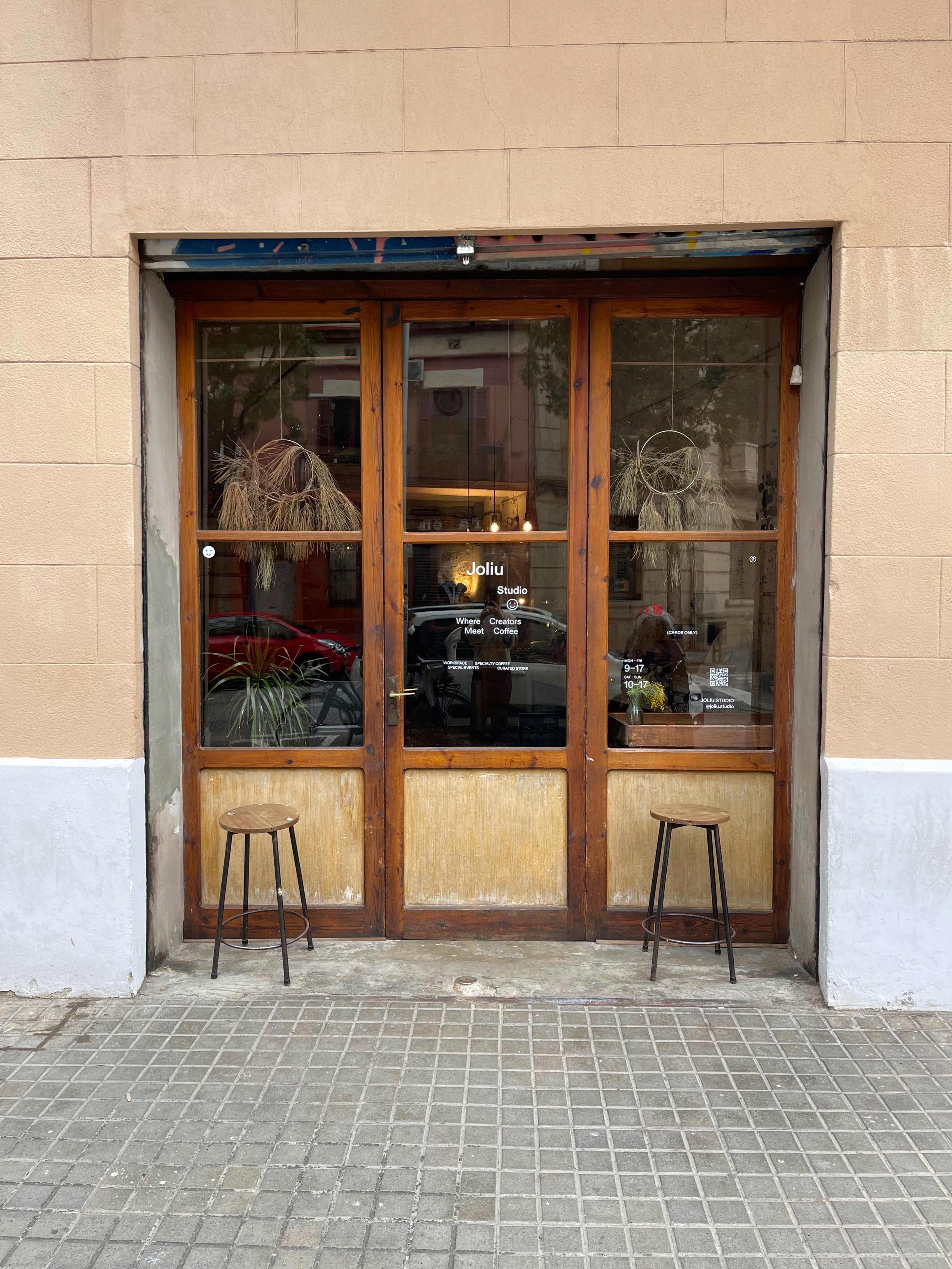 A wooden-framed storefront with large glass windows, two stools outside, and a sign that reads "Jolliu" on the door.