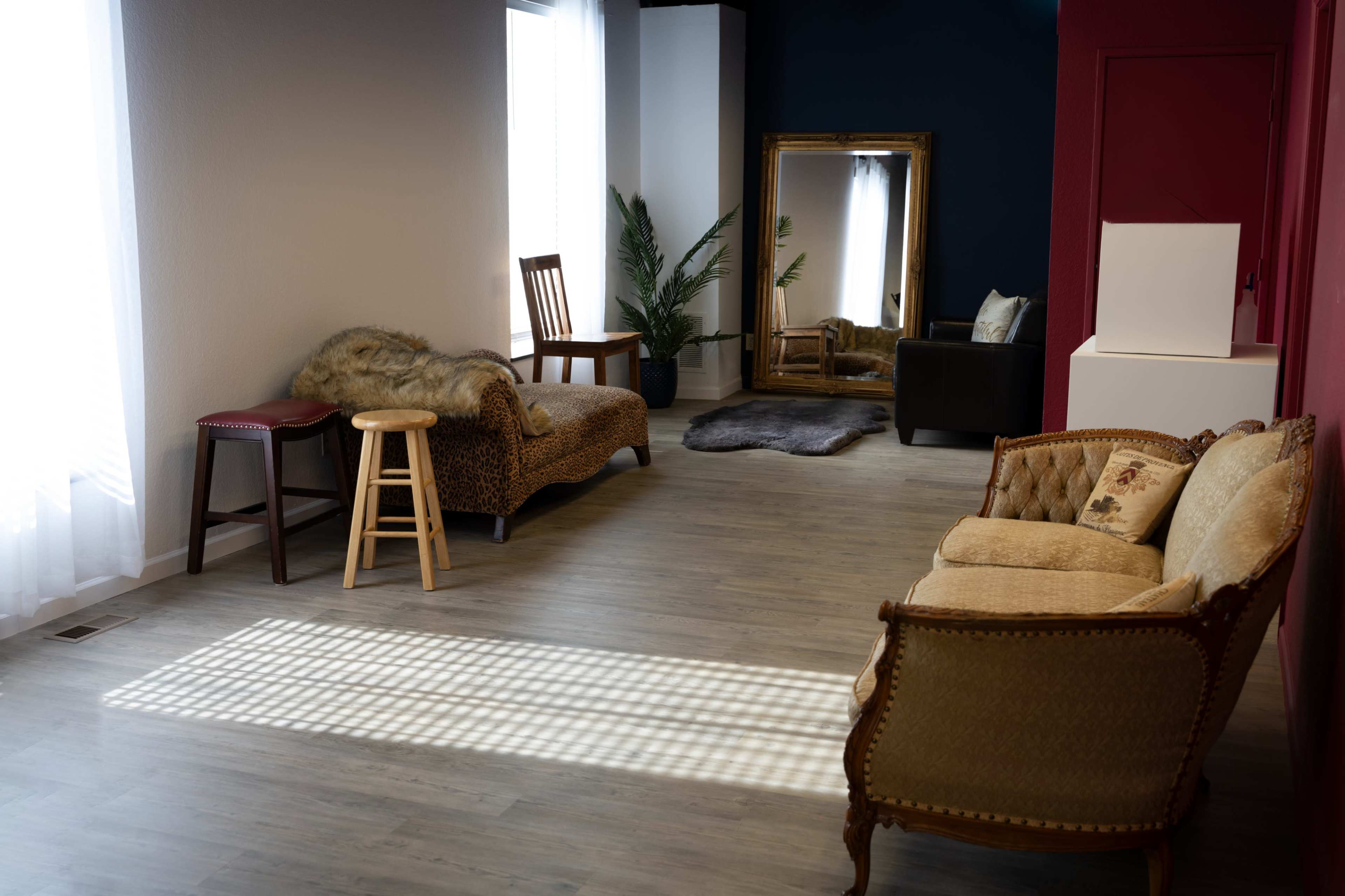 A spacious room with various seating arrangements, including a vintage couch, a wooden stool, and a chaise lounge, along with a large mirror and a potted plant.