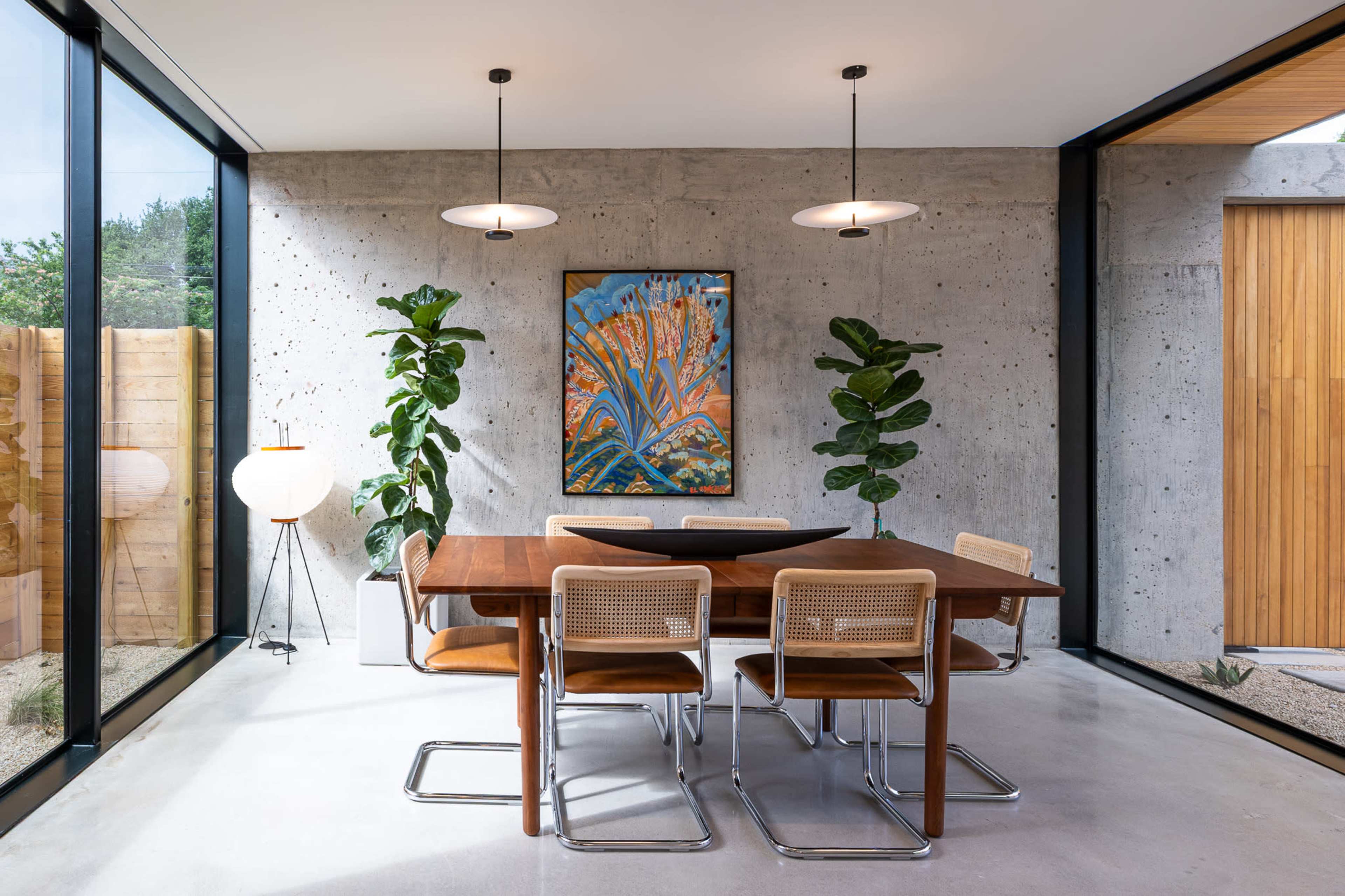 A modern dining area features a wooden table with metal chairs, surrounded by large windows and concrete walls, illuminated by pendant lights.