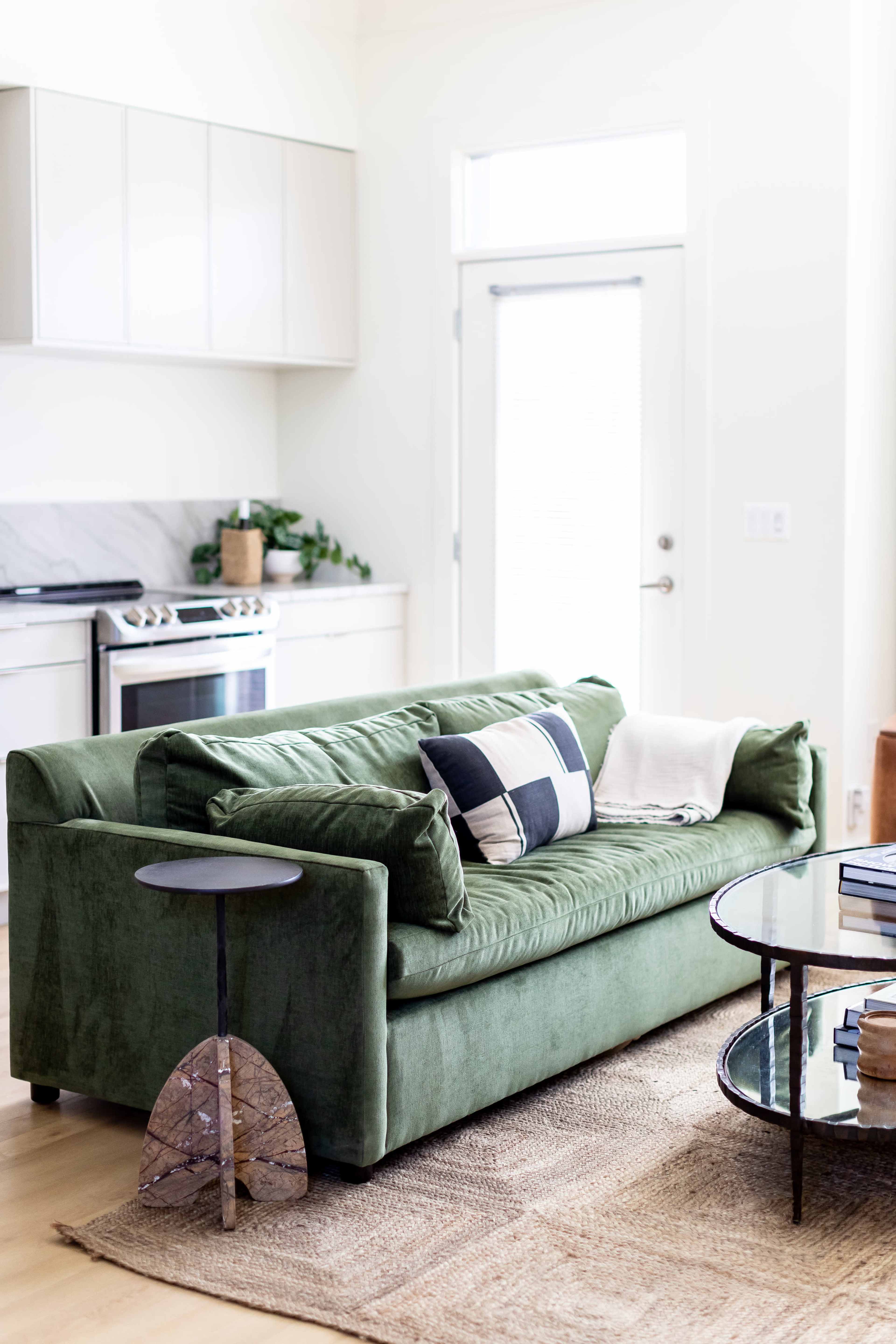 A green sofa with decorative pillows is positioned in a bright living room featuring a round coffee table and a kitchen in the background.