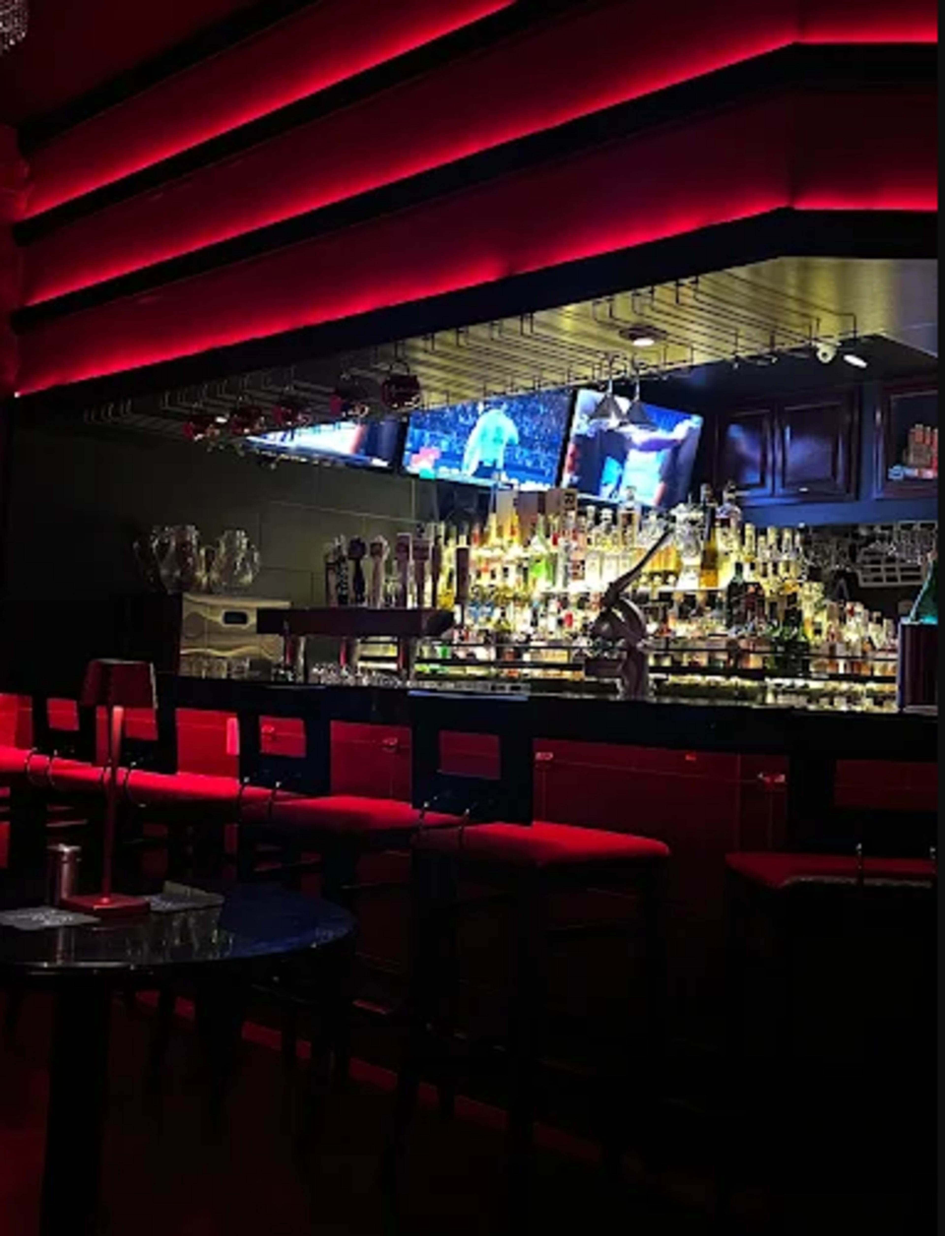 The image shows a dimly lit bar with red accents, featuring a well-stocked bar counter and multiple televisions displaying sports.
