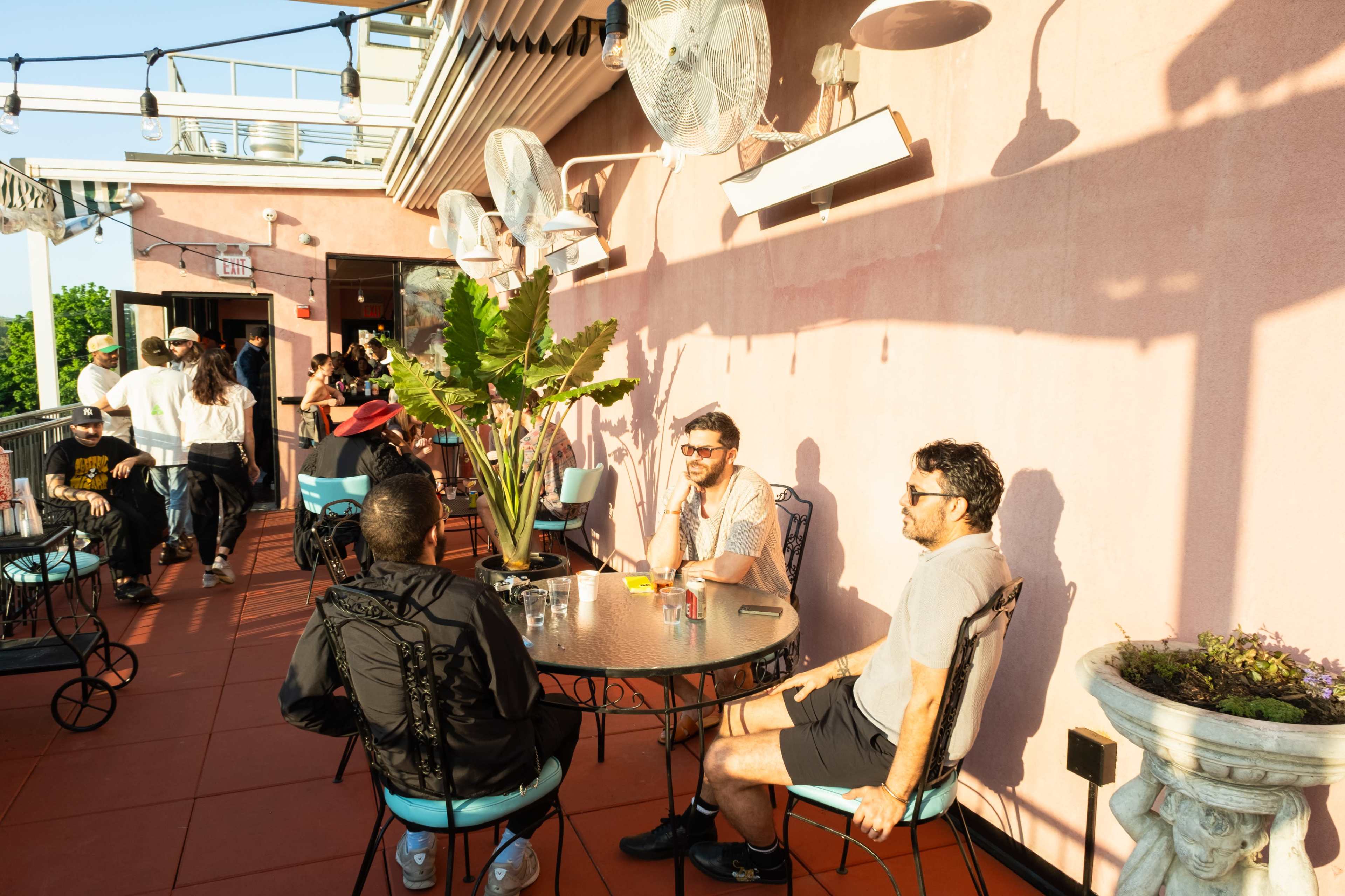 A group of people sits at a table on a patio, surrounded by plants and fans, with a pink wall in the background.