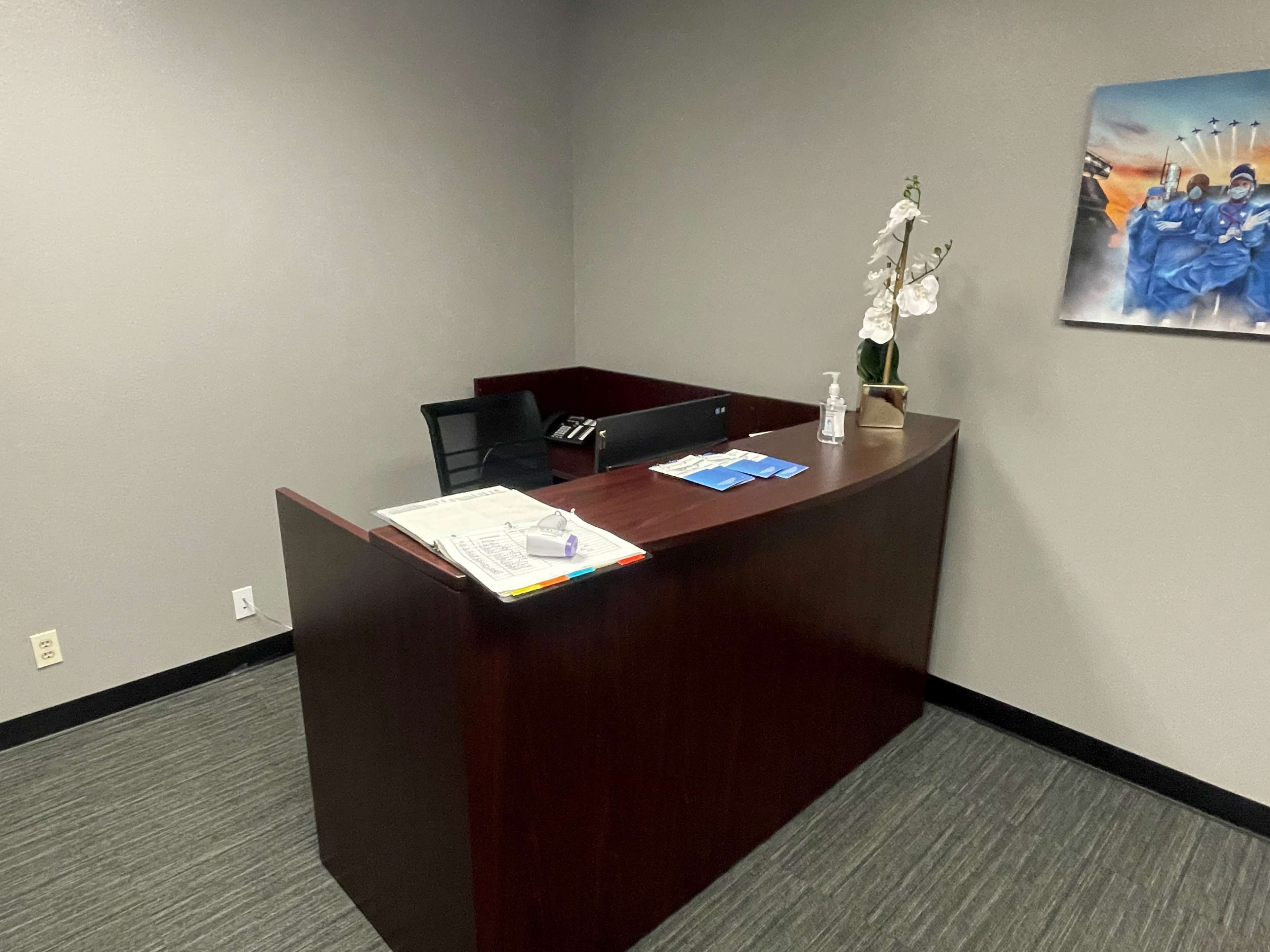 A modern office reception area with a wooden desk, a chair, and a floral decoration, set against a gray wall.