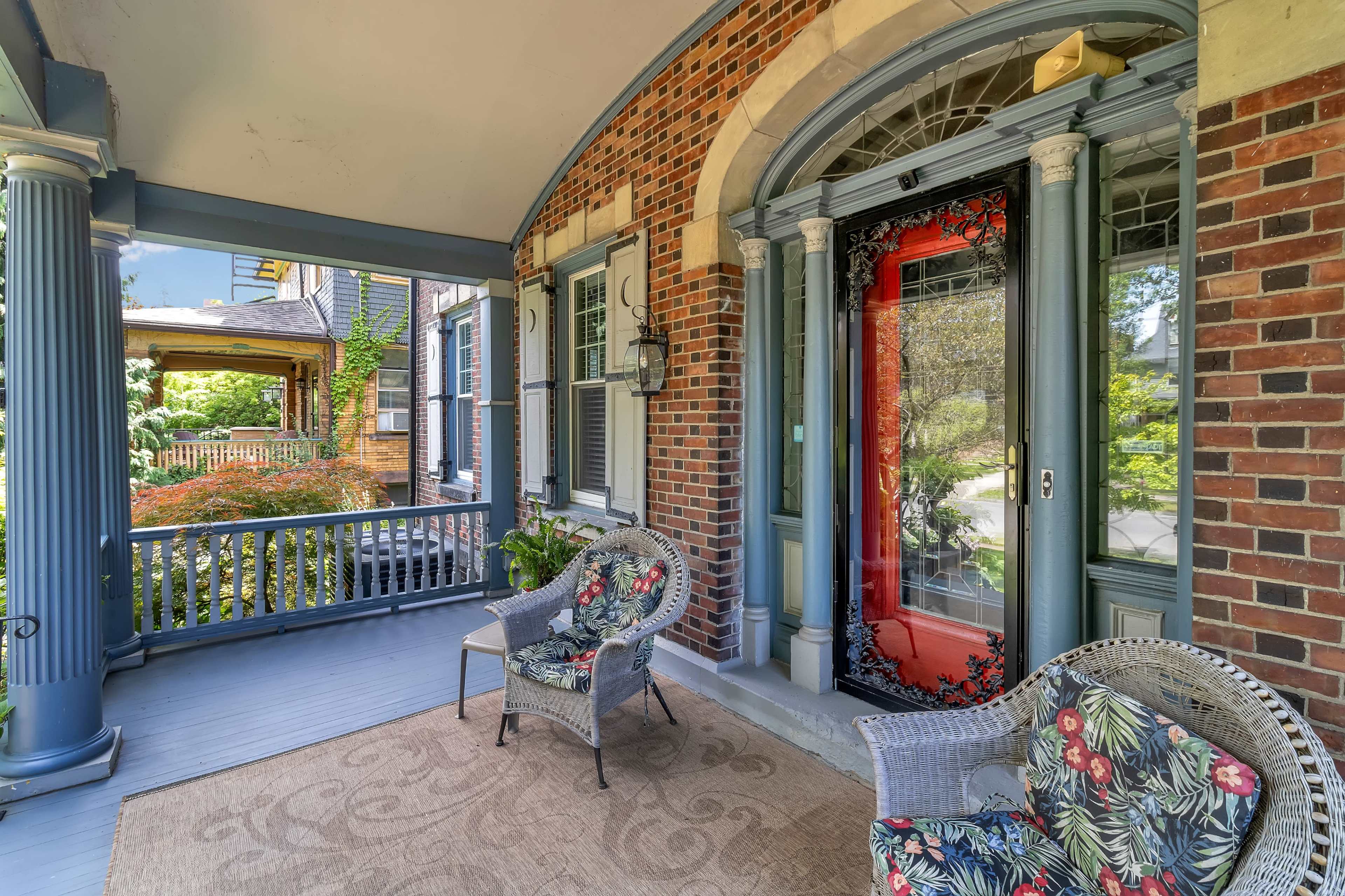 A porch featuring two floral-patterned chairs, a woven rug, and a bright red front door framed by decorative columns.