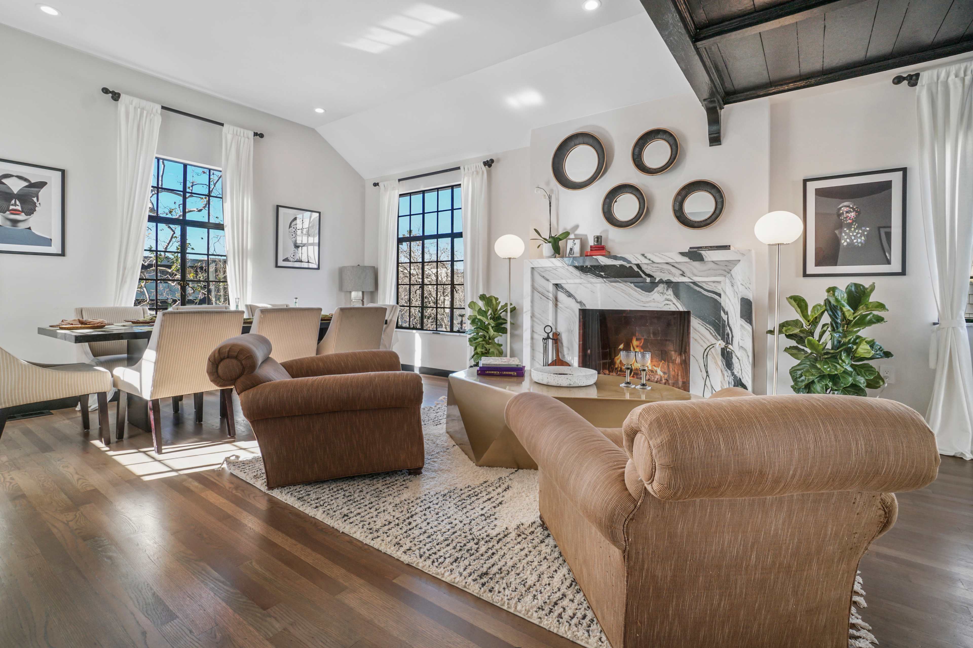 A modern living room features a marble fireplace, a coffee table, two armchairs, and large windows with natural light.