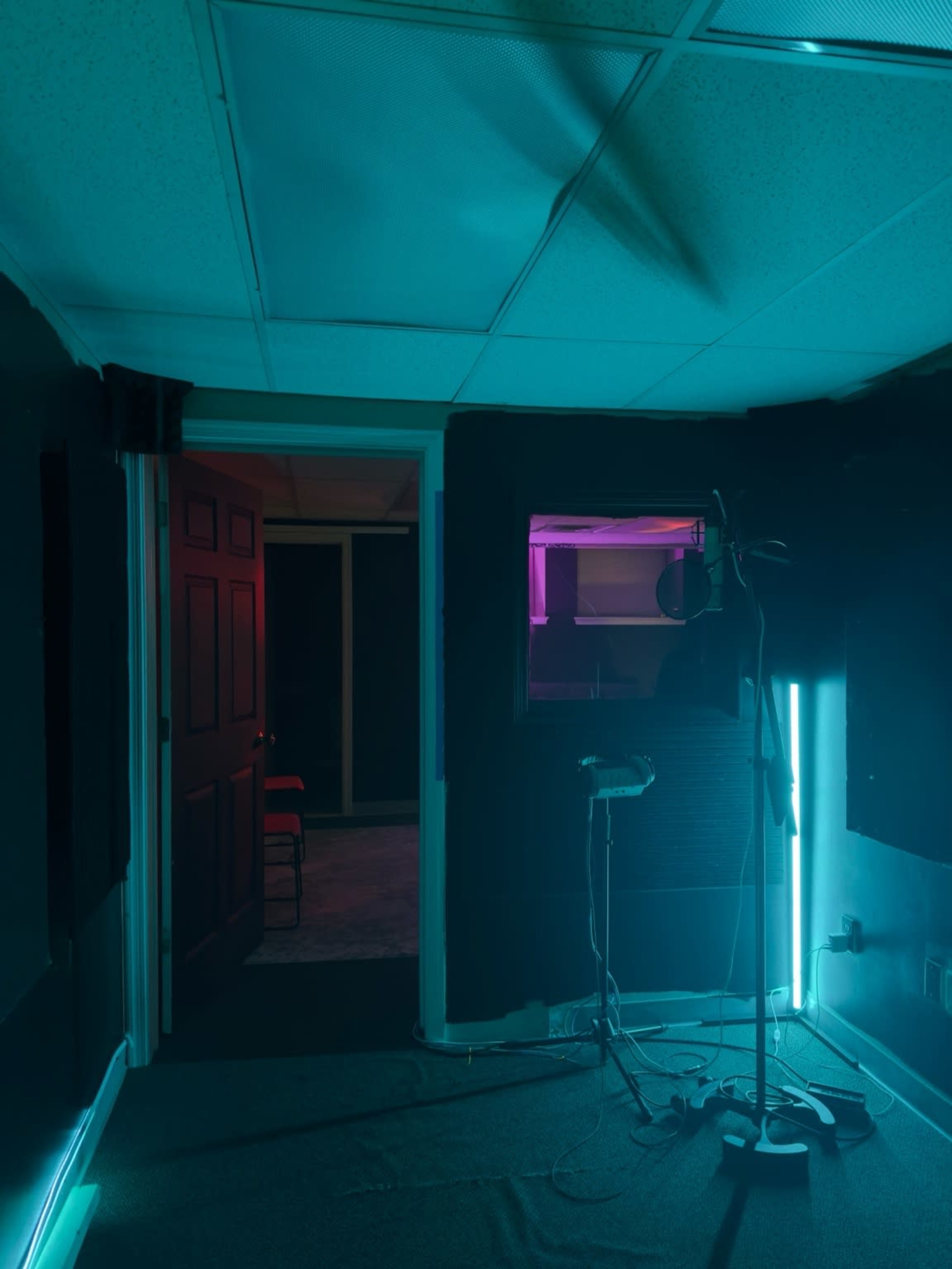A sound studio features a microphone on a stand, illuminated by blue and purple lights, with an open door revealing another room.