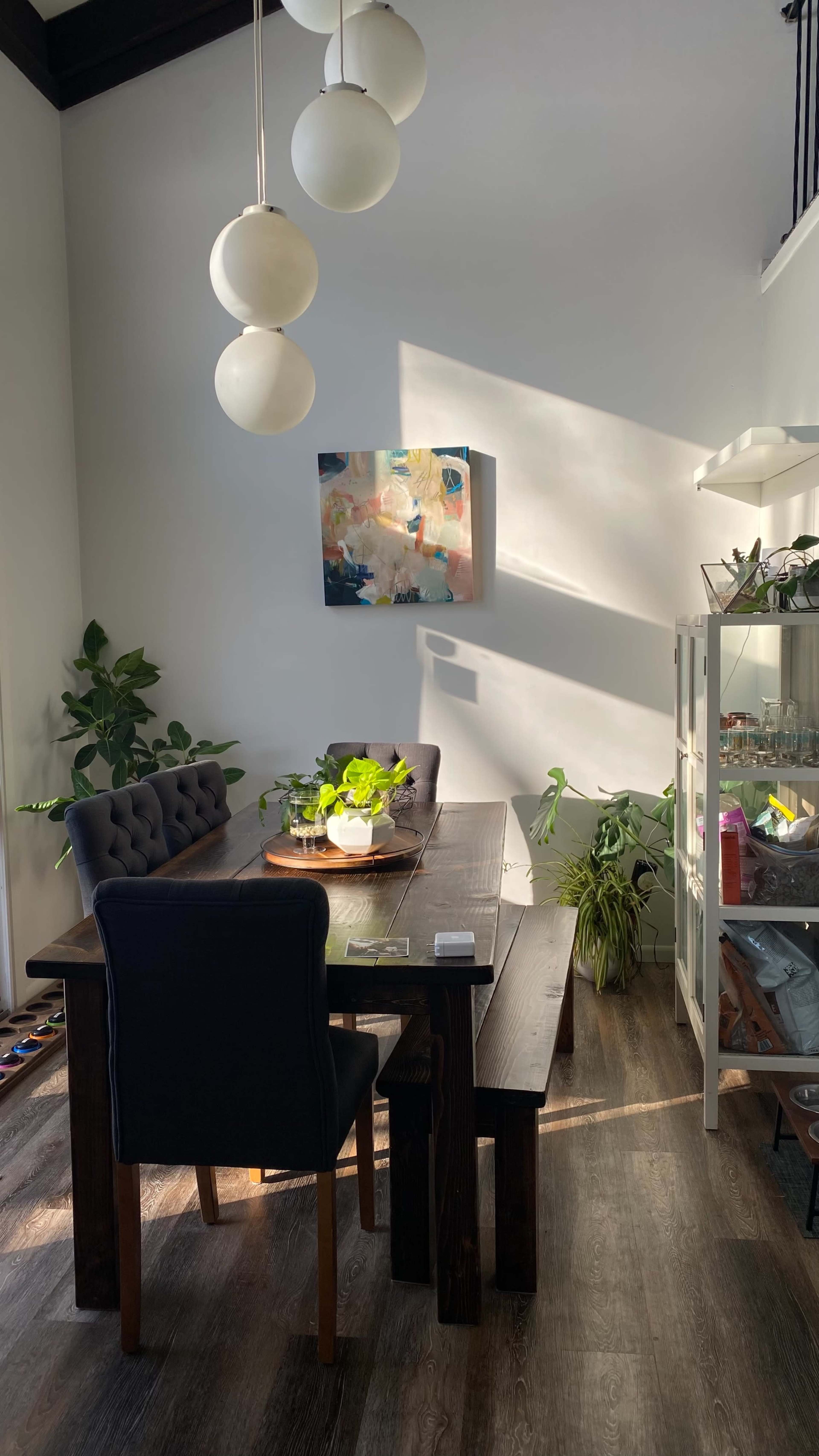 A dining area features a long wooden table surrounded by upholstered chairs, with a large painting on the wall and several potted plants nearby.