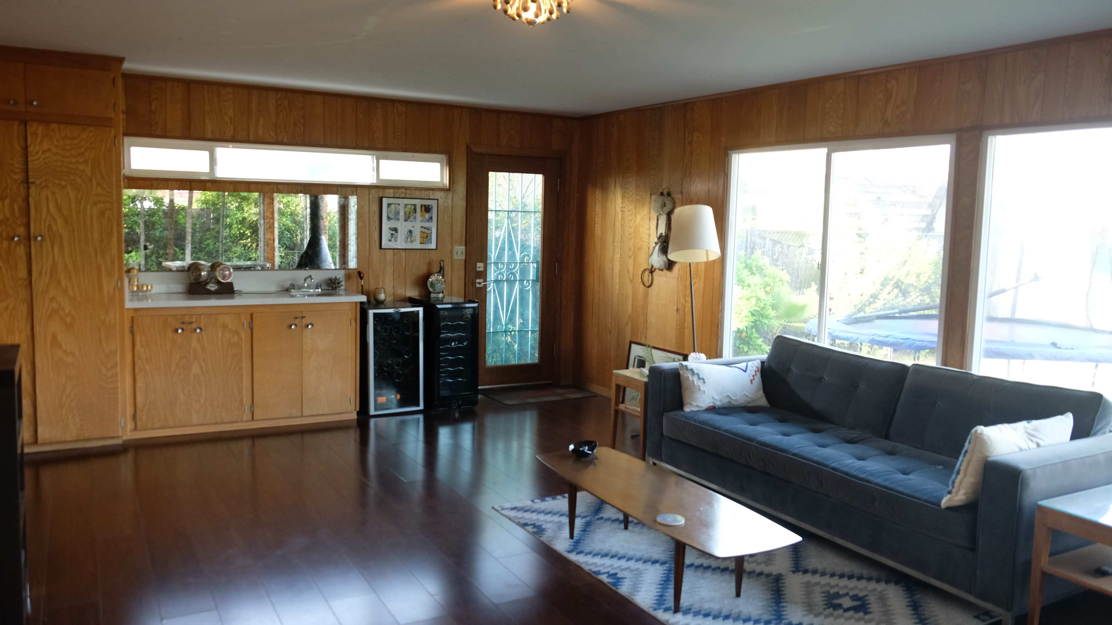 The image shows a living room with wooden walls, a large window, a black couch, a wooden coffee table, and a kitchen area in the background.