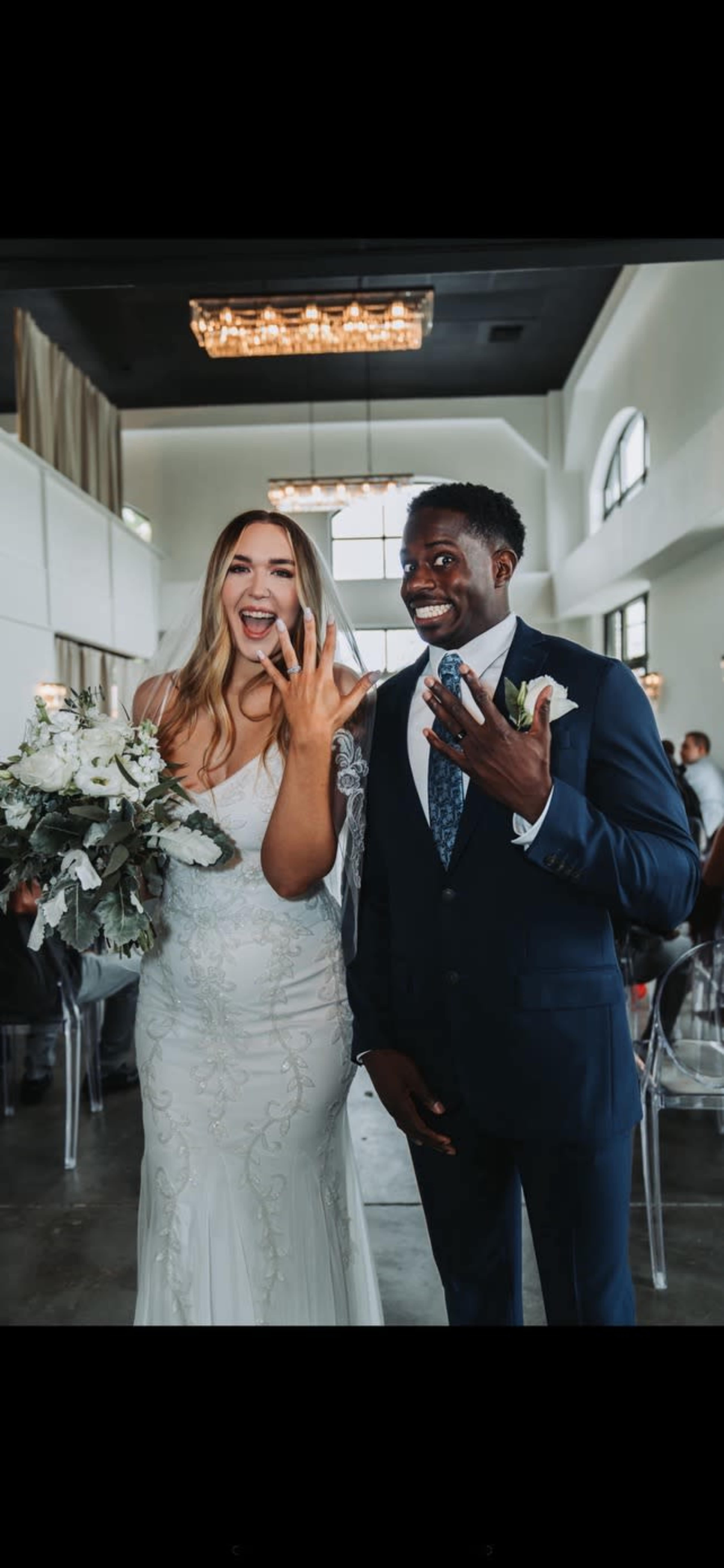 A bride and groom proudly display their wedding rings while smiling at the camera during their wedding reception.