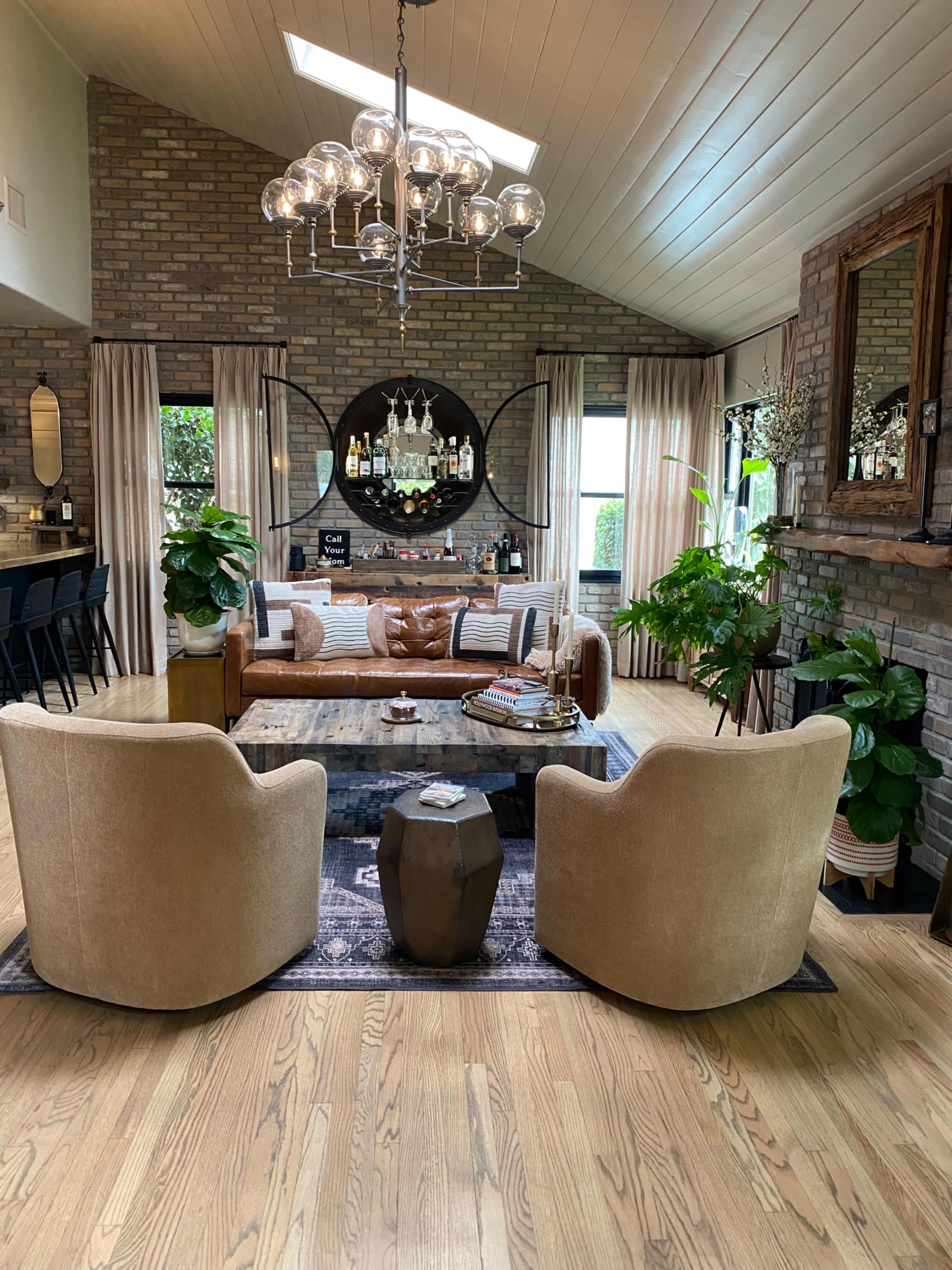 The image depicts a modern living room with a brown leather sofa, two beige chairs, a circular coffee table, and various decorative plants, all set against a brick wall with a stylish chandelier above.