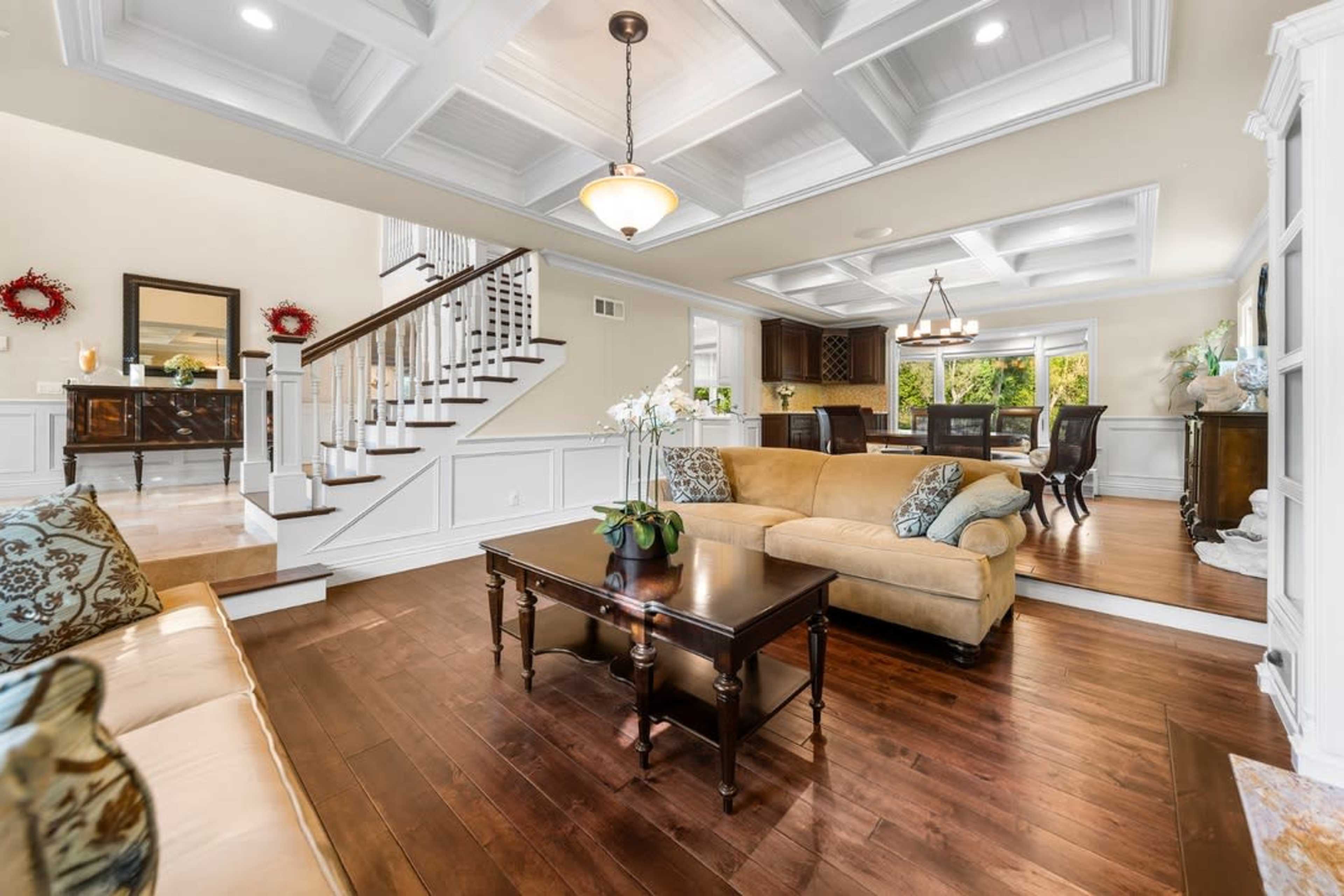 The image depicts a spacious living room with a coffered ceiling, a beige sofa, a wooden coffee table, and a staircase leading to an upper level.