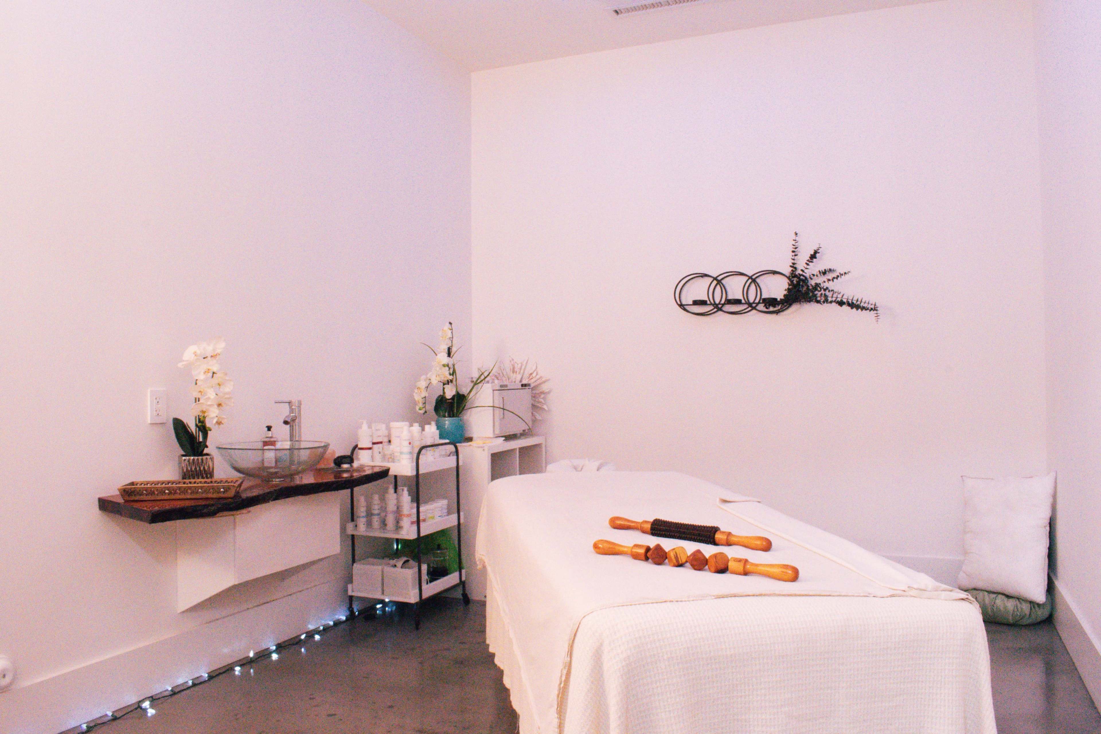 A massage room features a treatment table, light-colored walls, and a small shelf with skincare products and decorative items.