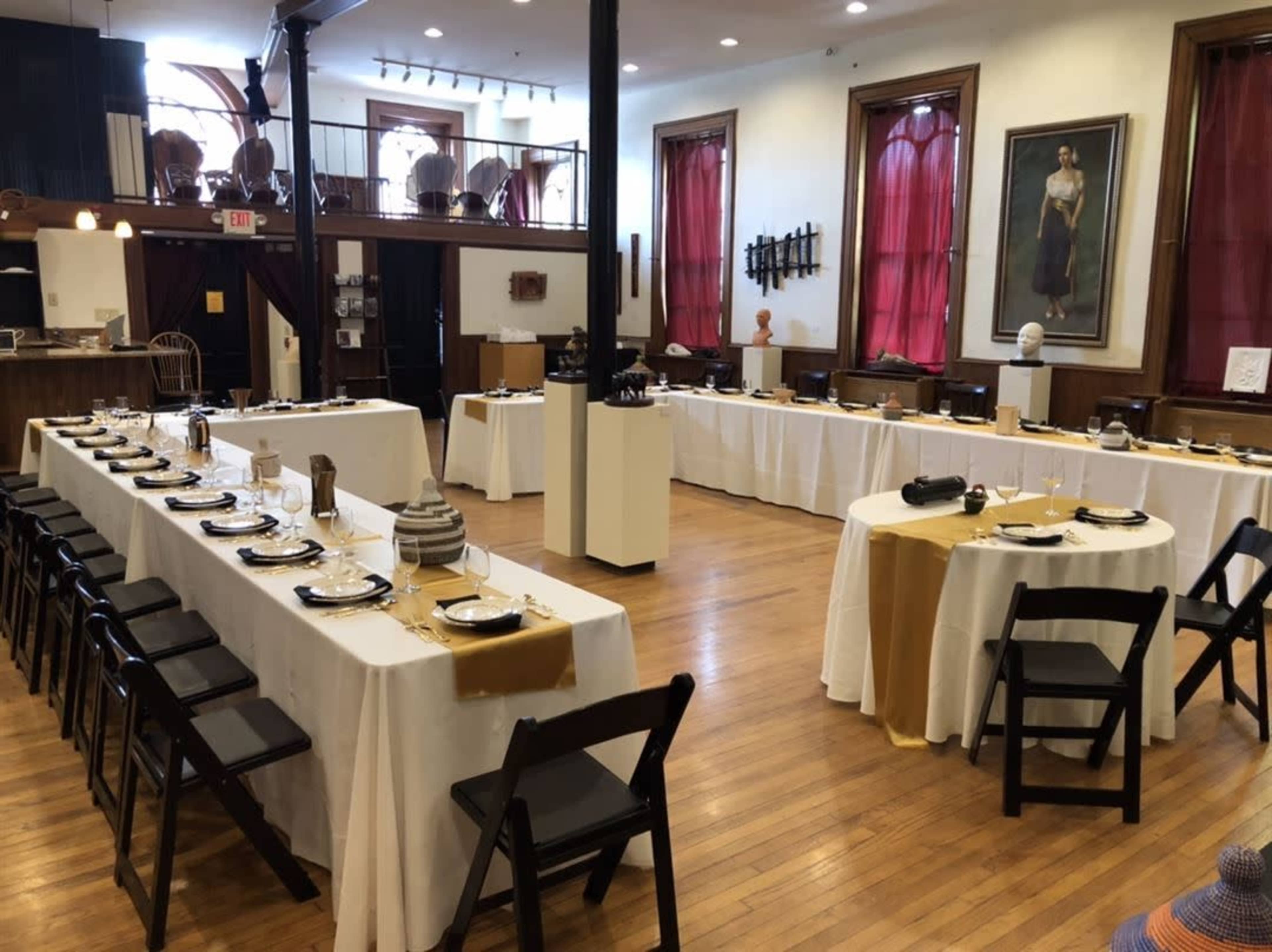 The image shows a dining area set up with several long tables, each covered in white tablecloths and arranged with dishes and silverware, within a room featuring wooden floors and decorative wall art.