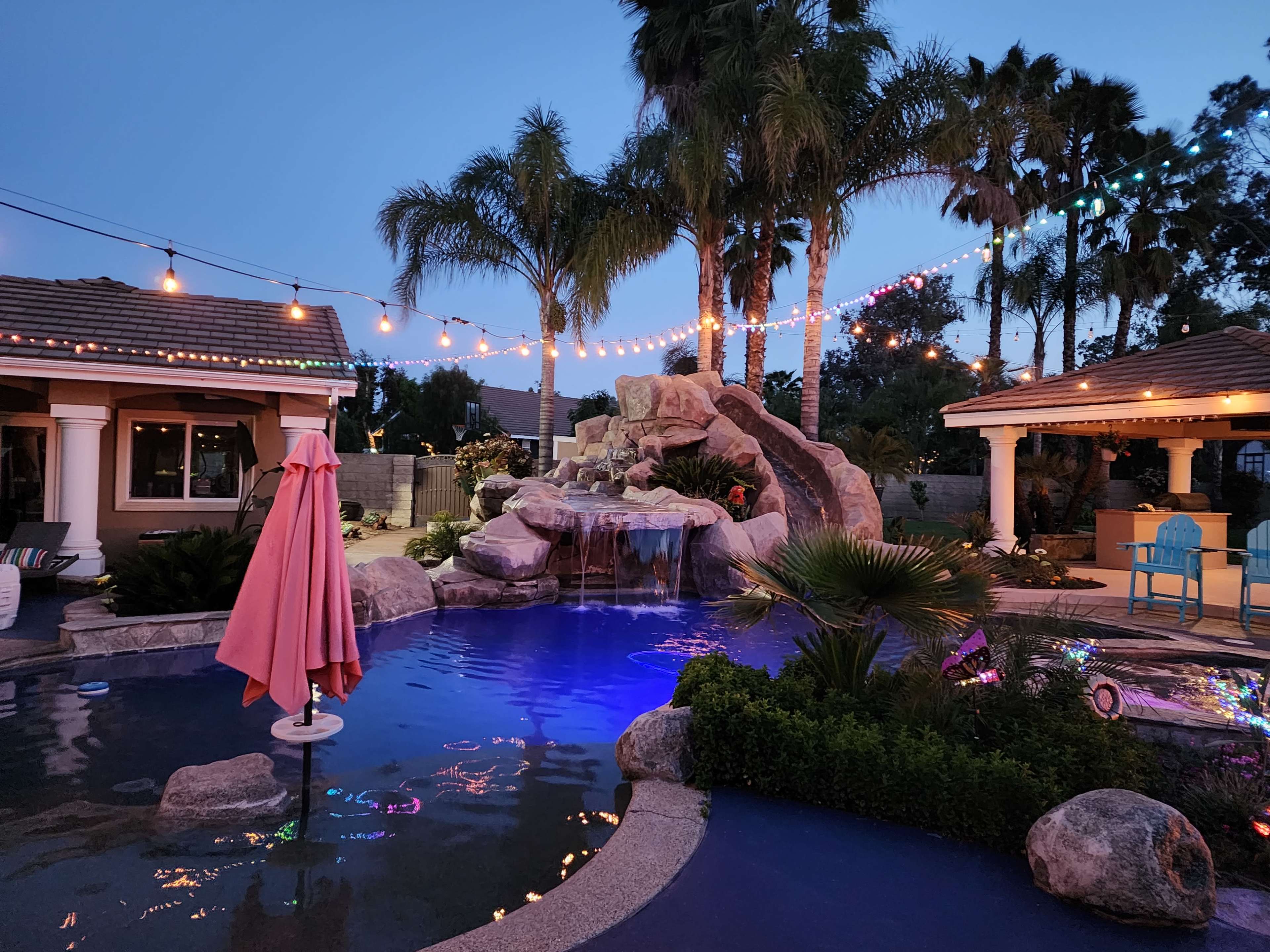 A swimming pool area with a rock waterfall, surrounded by palm trees and illuminated by string lights at dusk.
