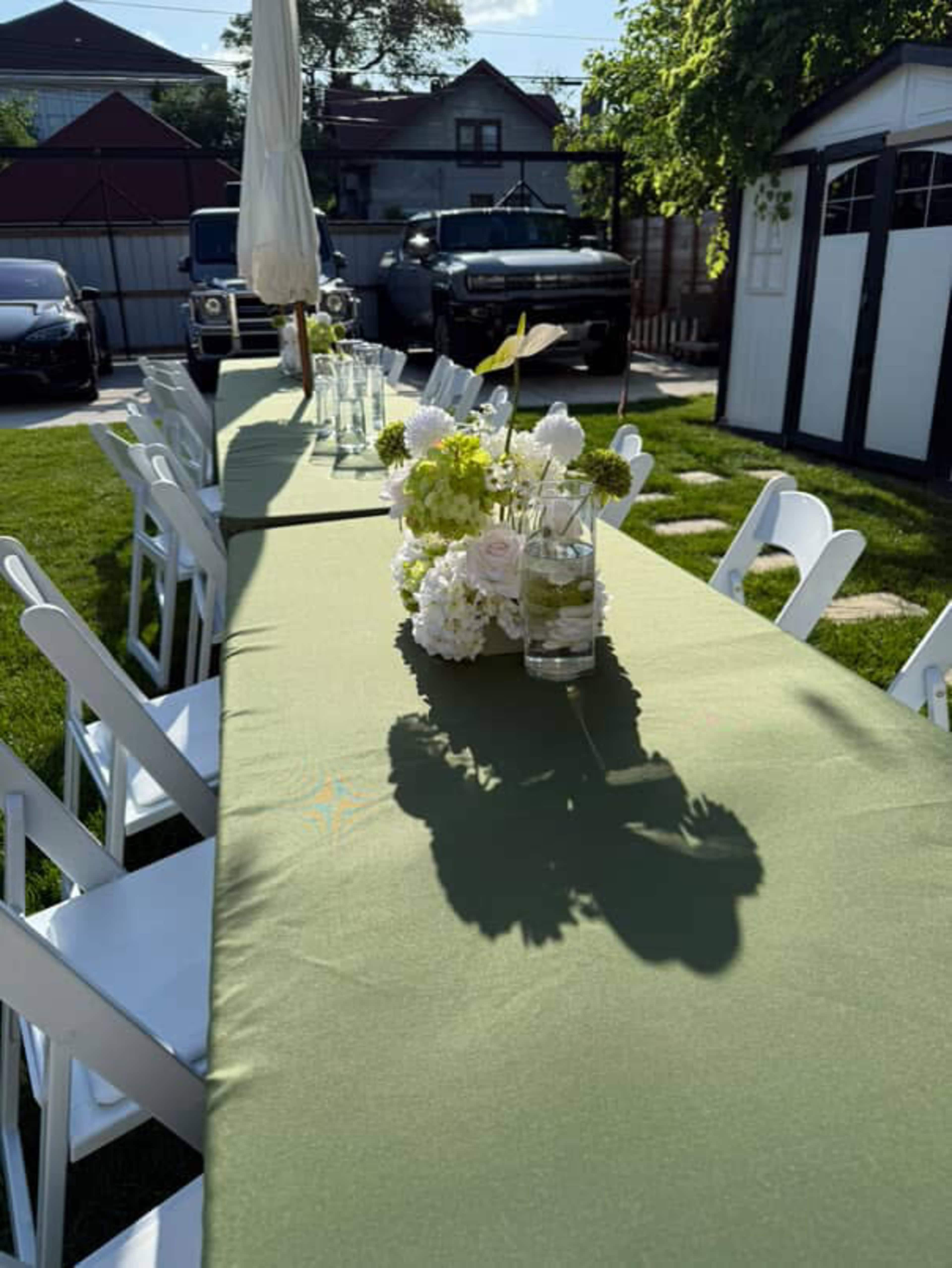 A long green table is set outdoors with white chairs, decorated with floral centerpieces and casting shadows in the sunlight.