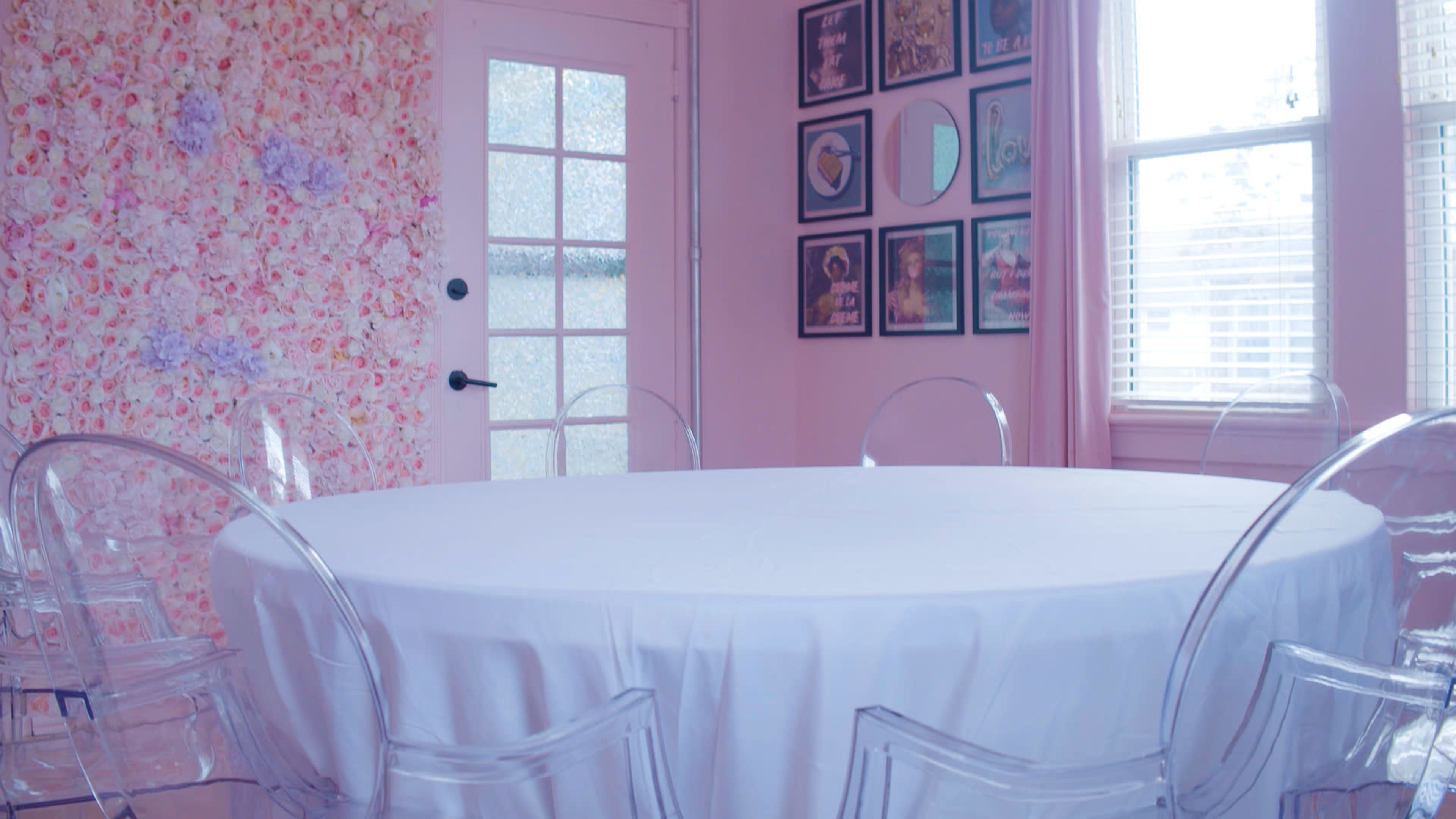 A round table covered with a white tablecloth is surrounded by clear acrylic chairs in a room with pink floral wallpaper and framed artwork on the walls.