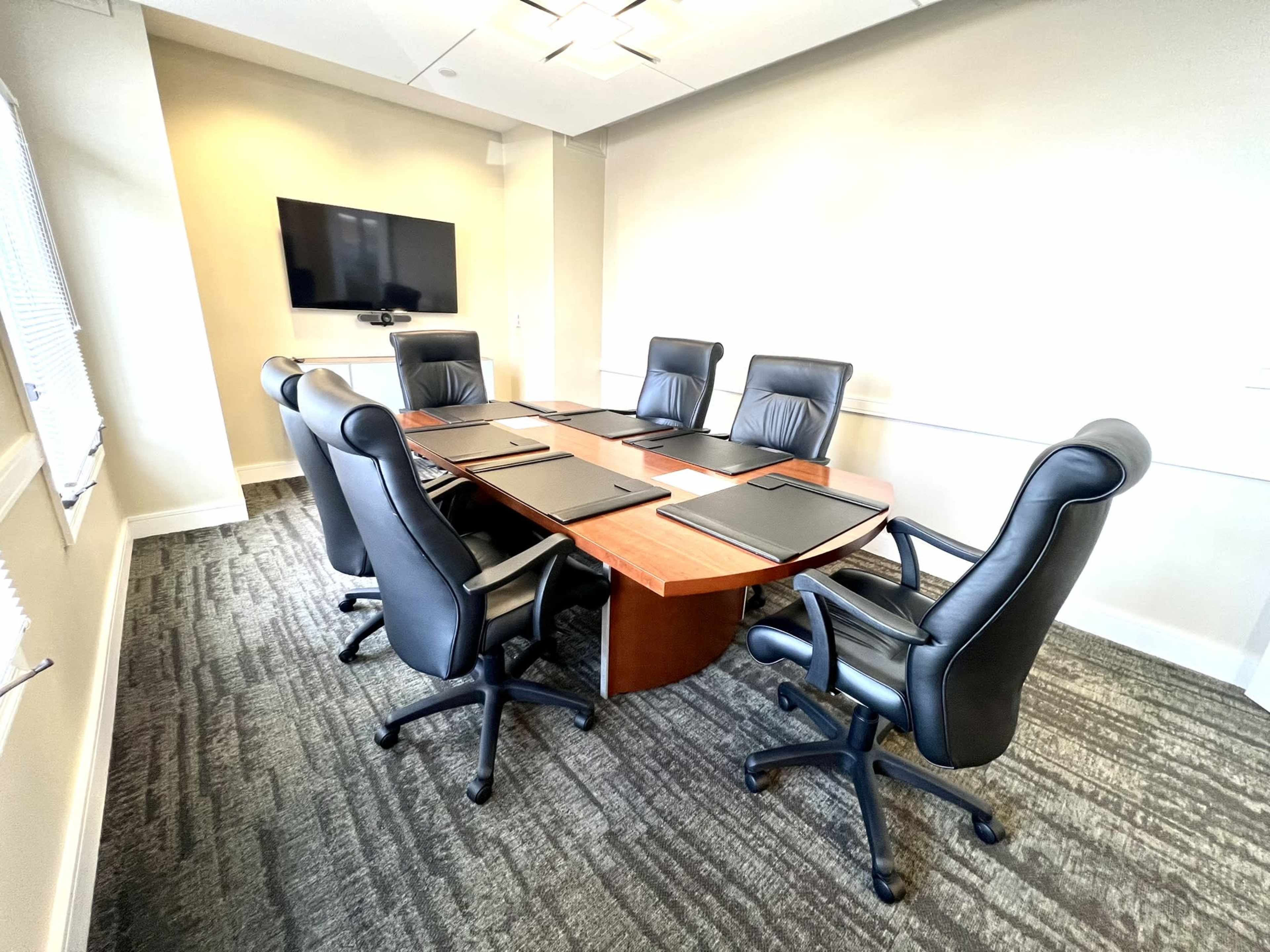 A conference room features a large oval table surrounded by six black leather chairs, with a television mounted on the wall.