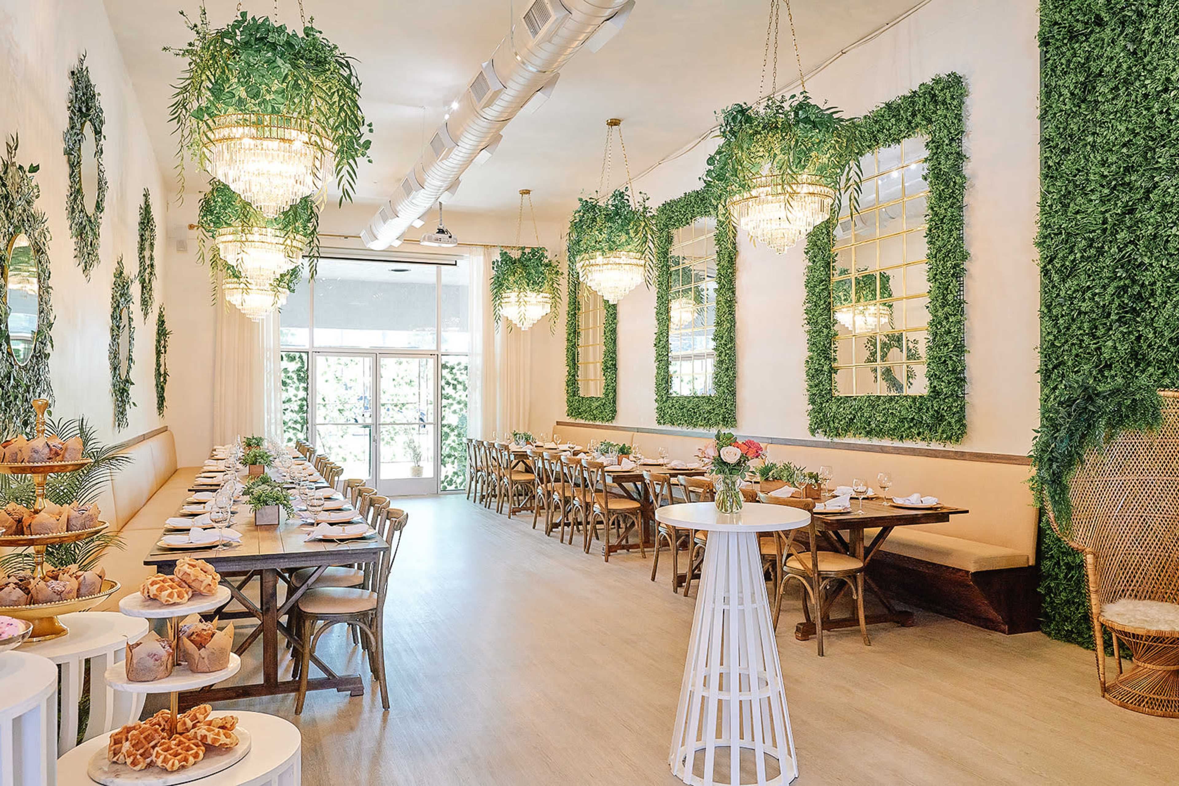The image shows a dining area with wooden tables, greenery on the walls, and large chandeliers hanging from the ceiling.