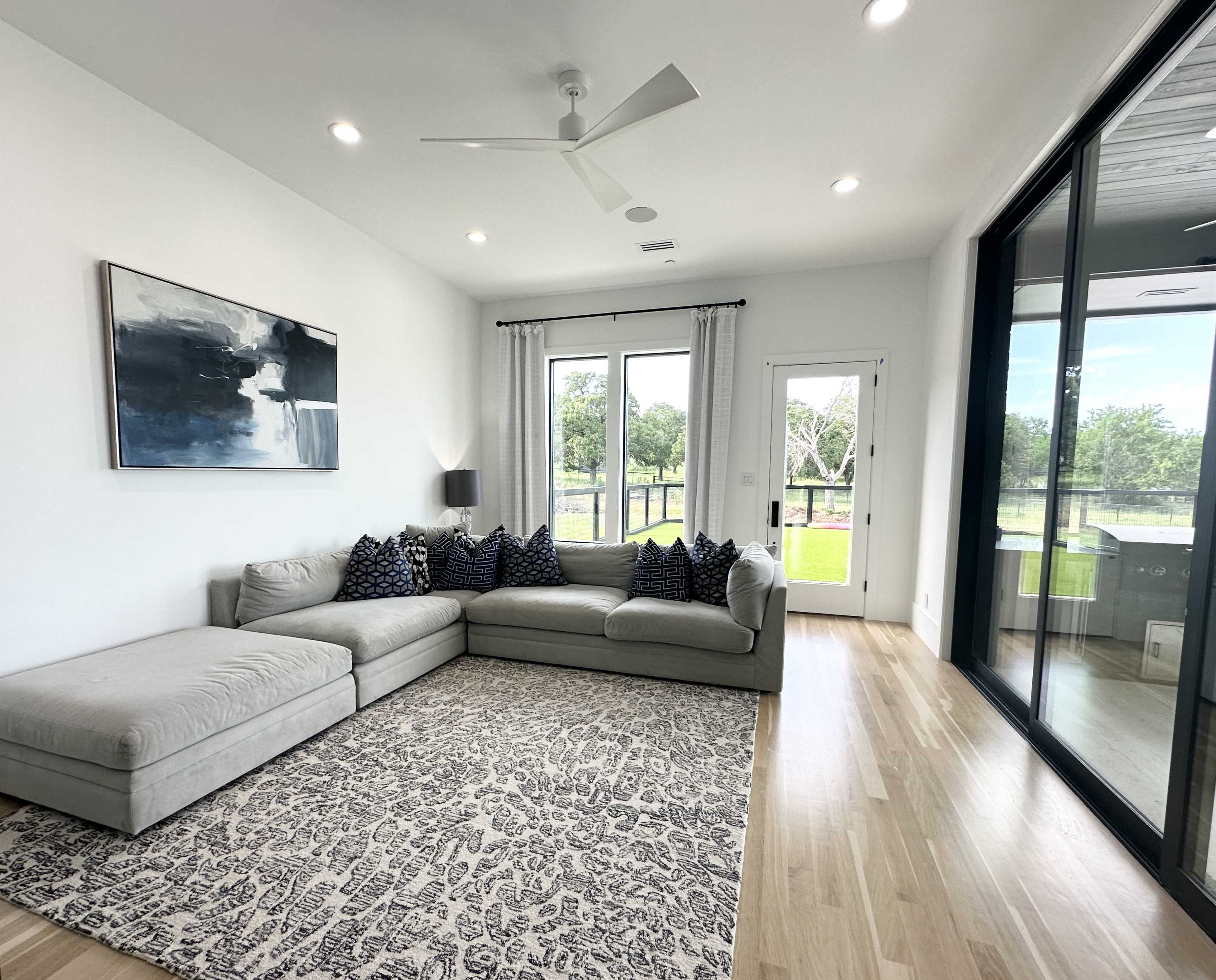 A spacious living room features a gray sectional sofa with patterned cushions, a large area rug, and floor-to-ceiling windows overlooking a green outdoor space.