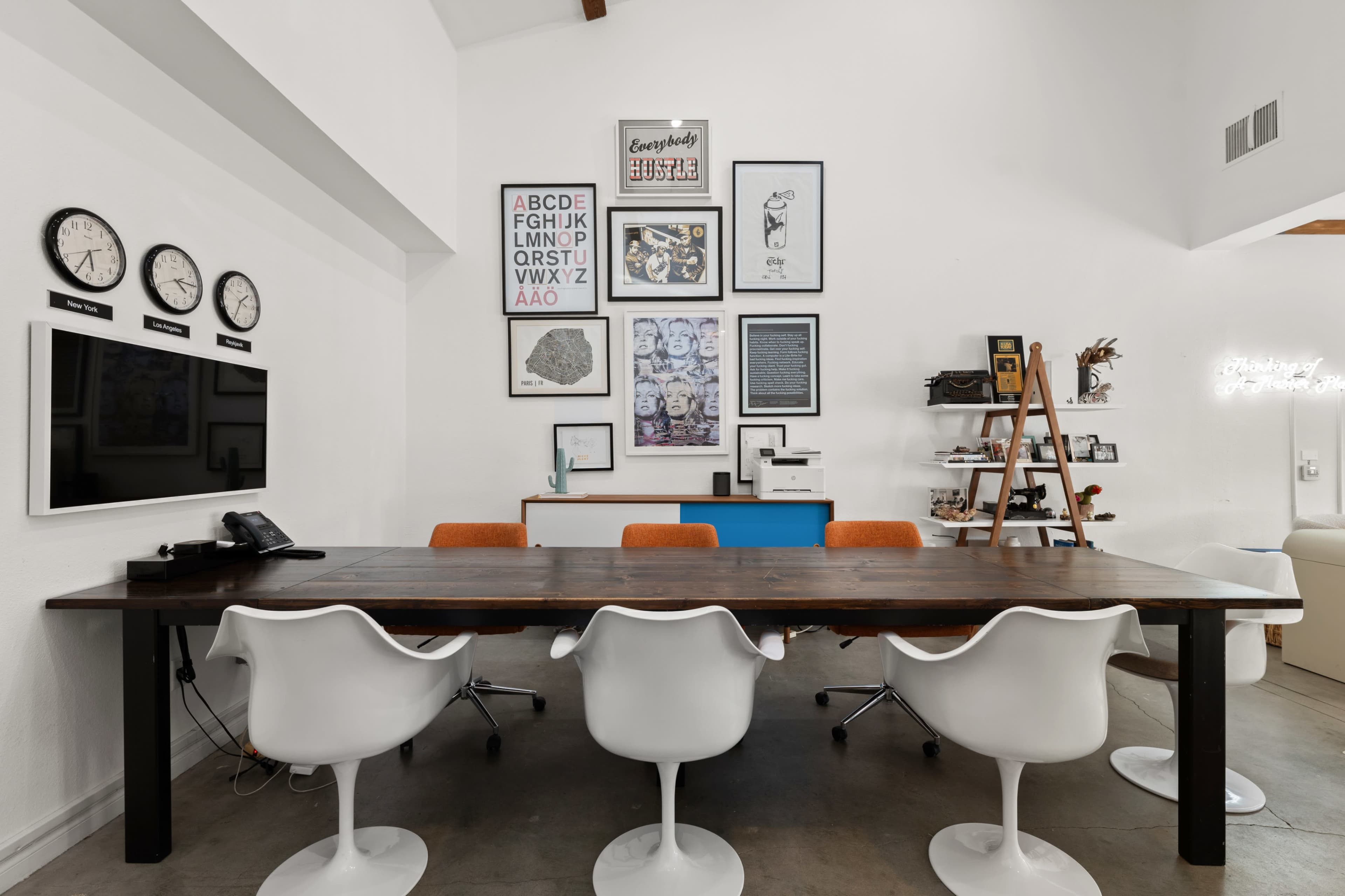 A modern meeting room features a long wooden table surrounded by white chairs, with wall art, clocks, and a console in the background.
