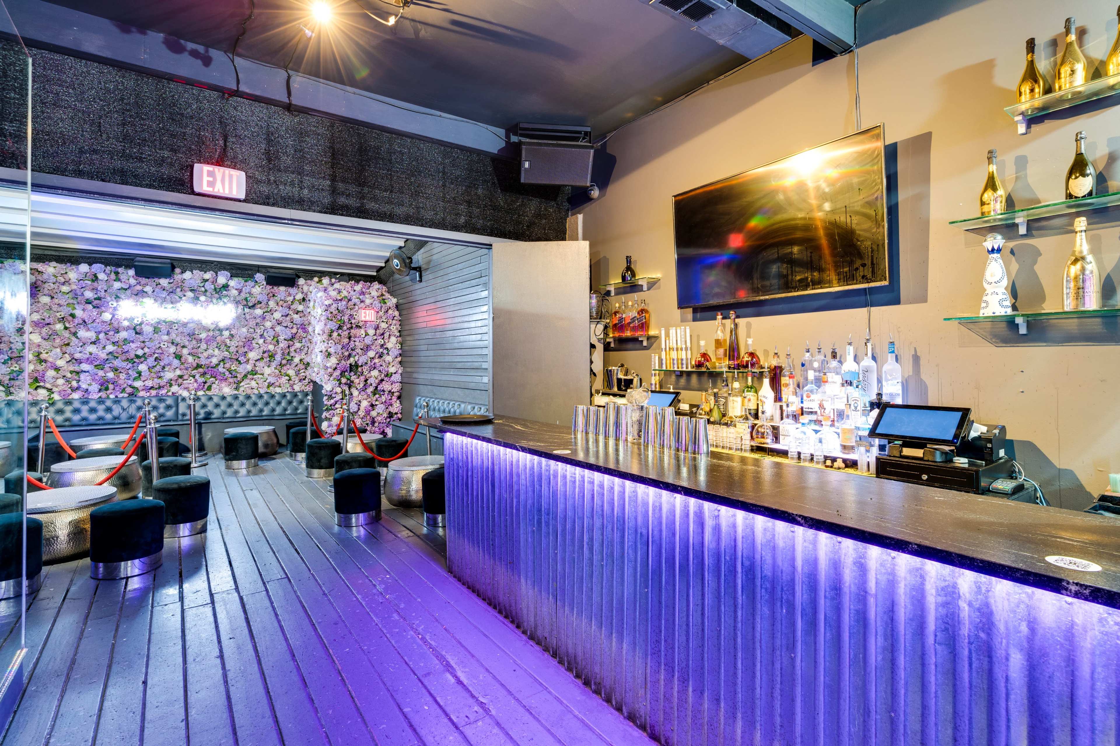 A bar interior with a purple-lit counter, a wall decorated with flowers, and seating arrangements on a wooden floor.