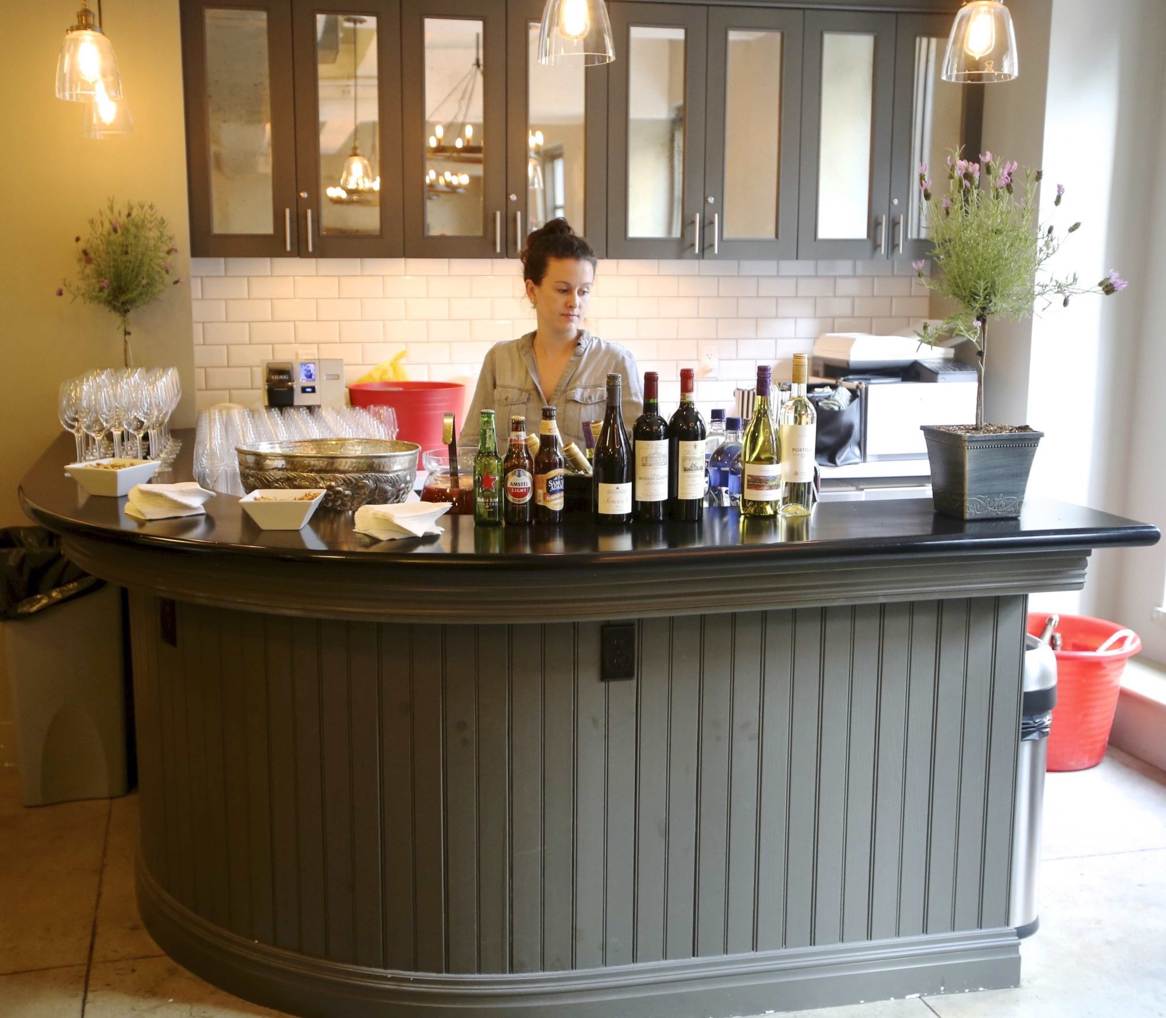 A woman stands behind a curved bar filled with various bottles of wine and beer, alongside glassware and plates.
