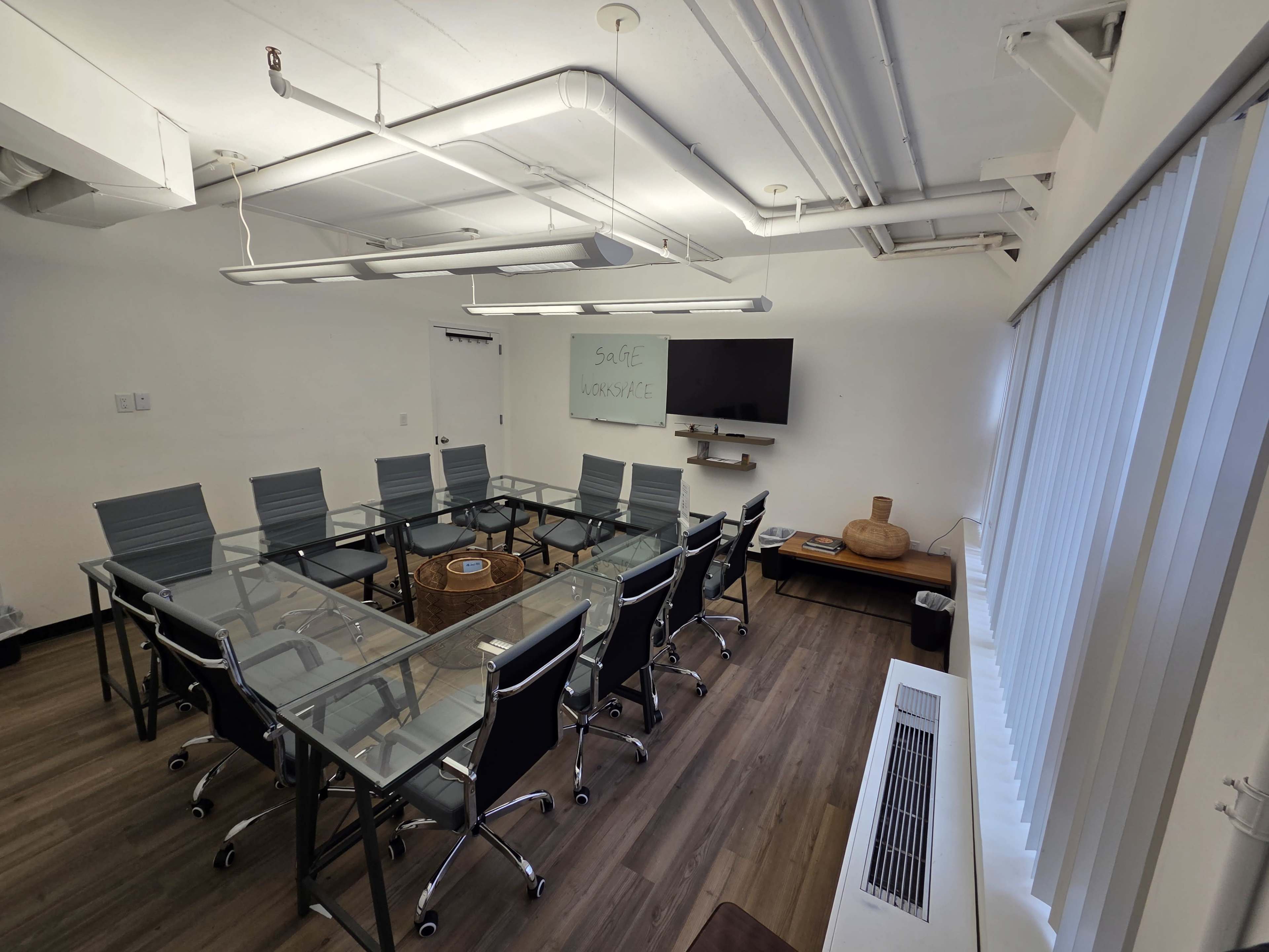 The image shows a modern conference room featuring a large glass table surrounded by eight office chairs, a flat-screen TV on the wall, and a large window with vertical blinds.