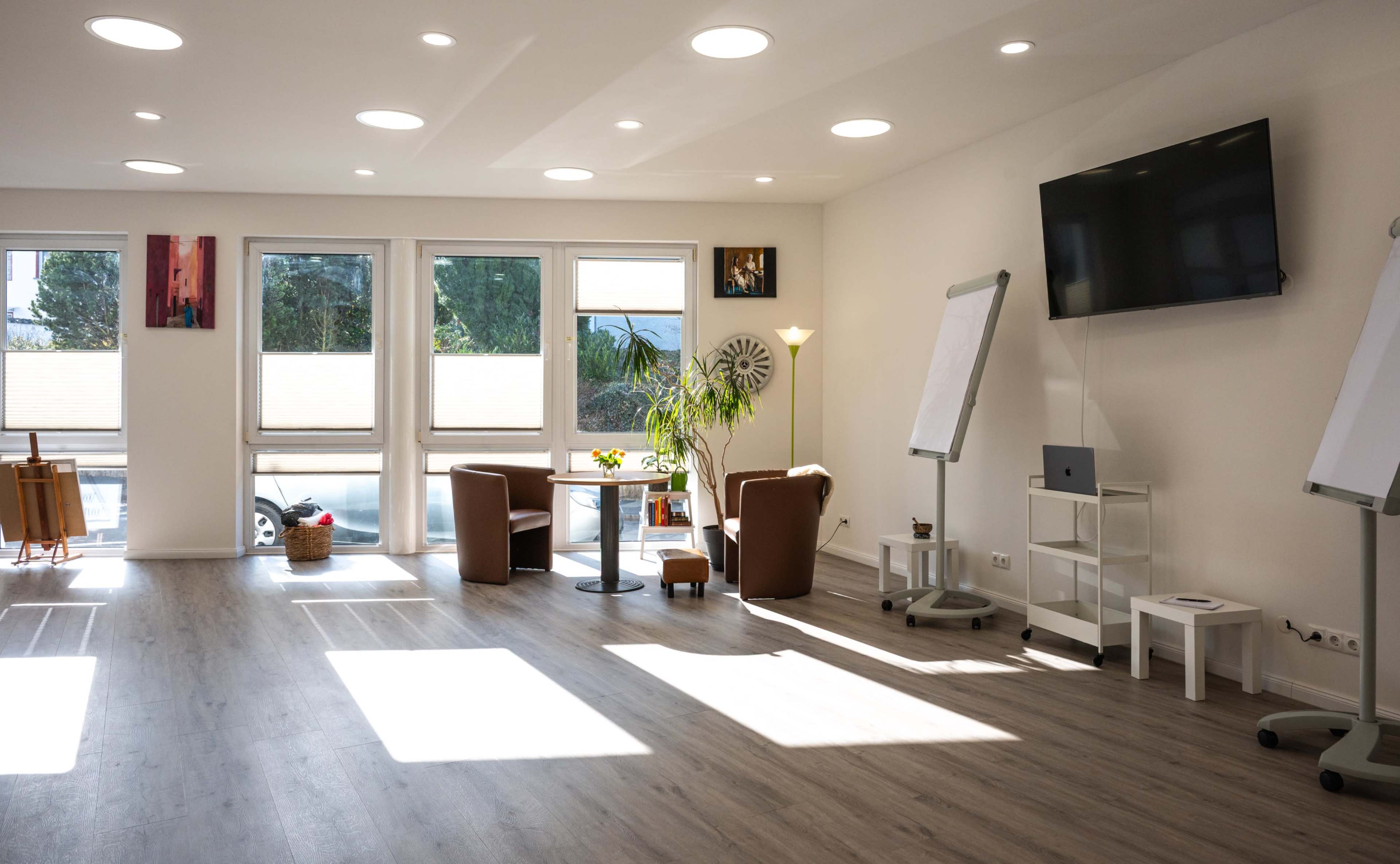 A bright, spacious room with large windows, wooden flooring, and seating areas, furnished with a TV, a table, and whiteboards.