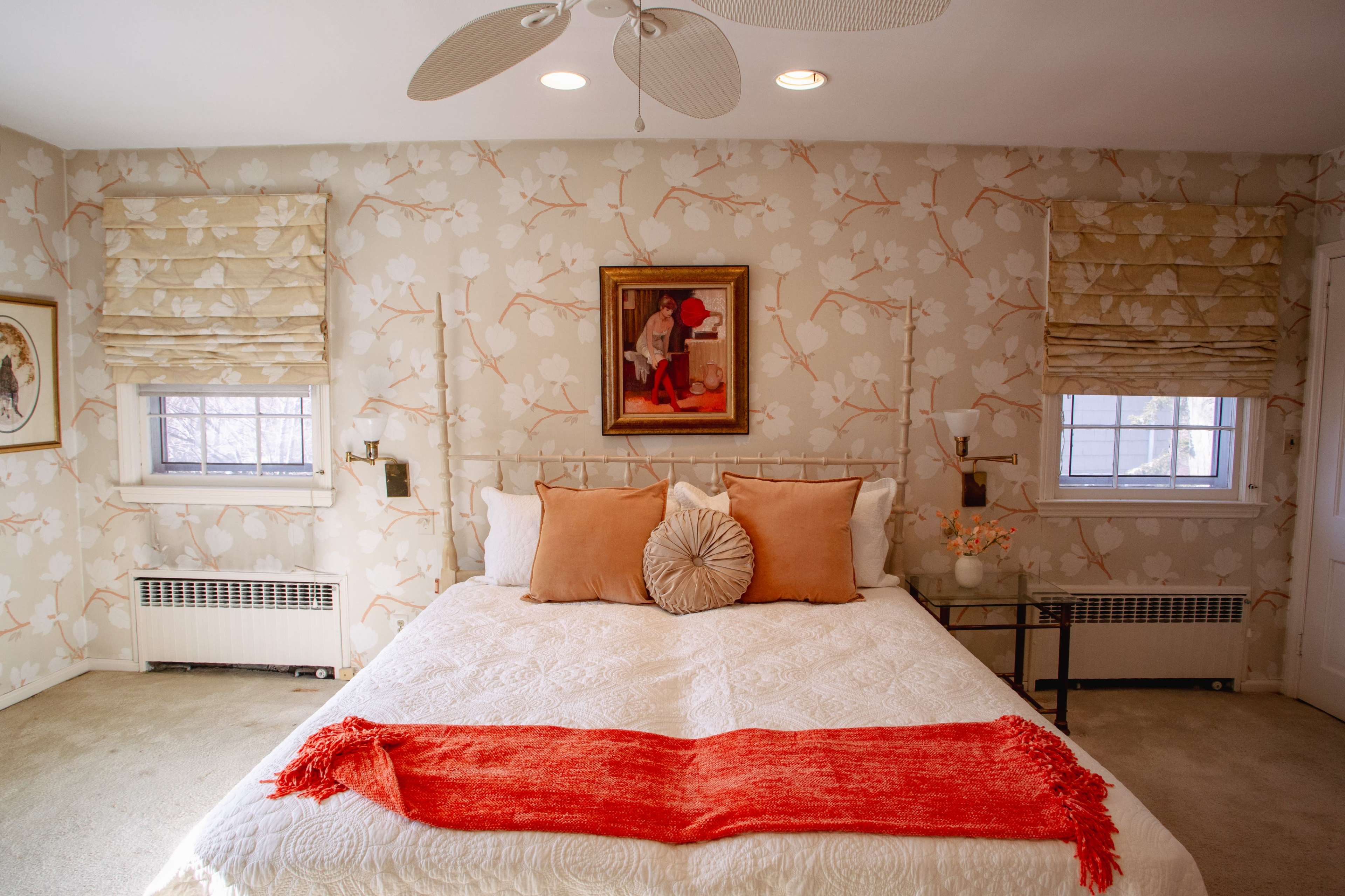 A neatly arranged bedroom features a bed with decorative pillows and a bright red throw blanket, surrounded by floral wallpaper and adorned with framed artwork.