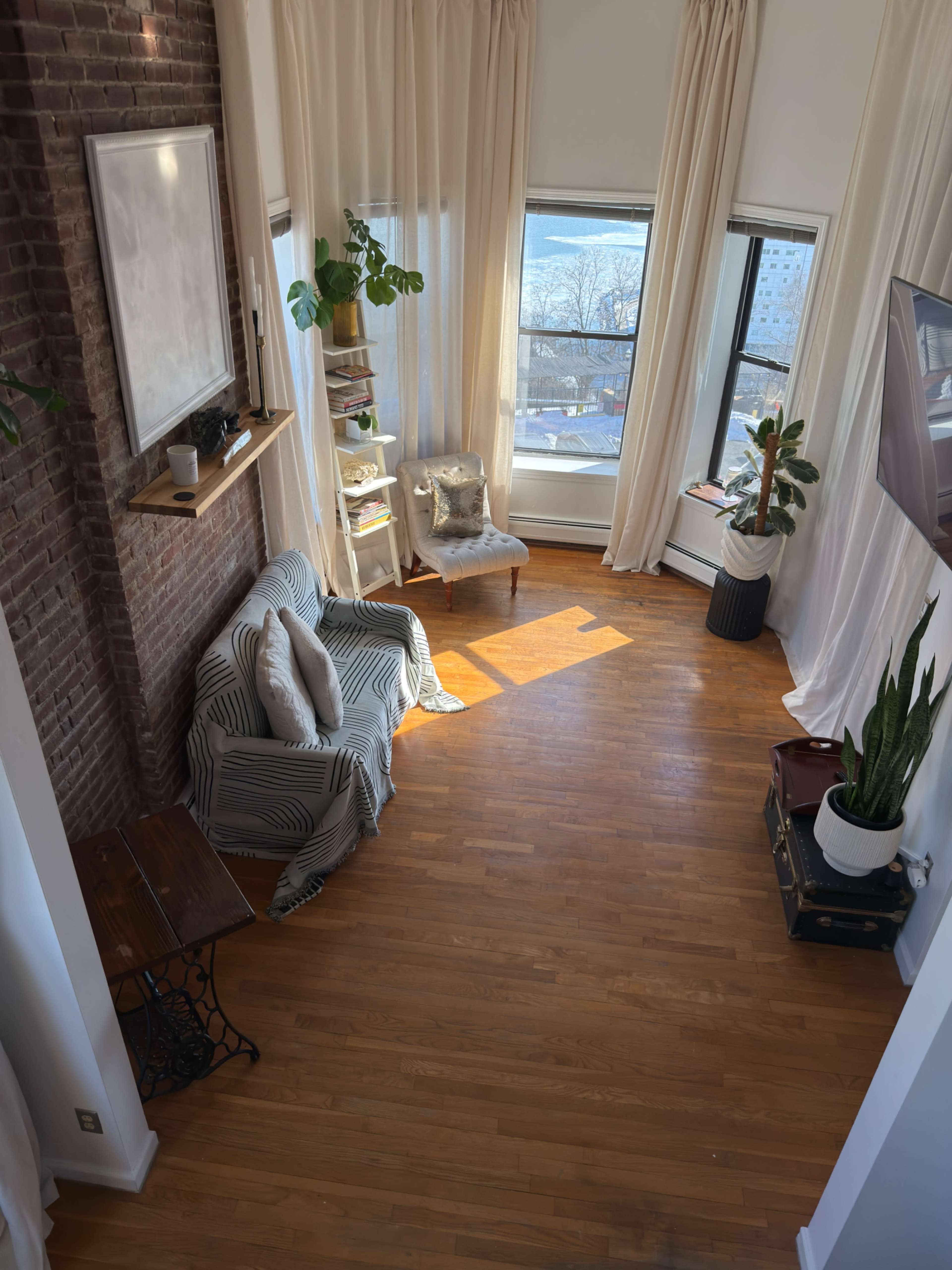 The image shows a bright, airy living room with large windows, hardwood flooring, a comfortable chair, plants, and a brick wall.