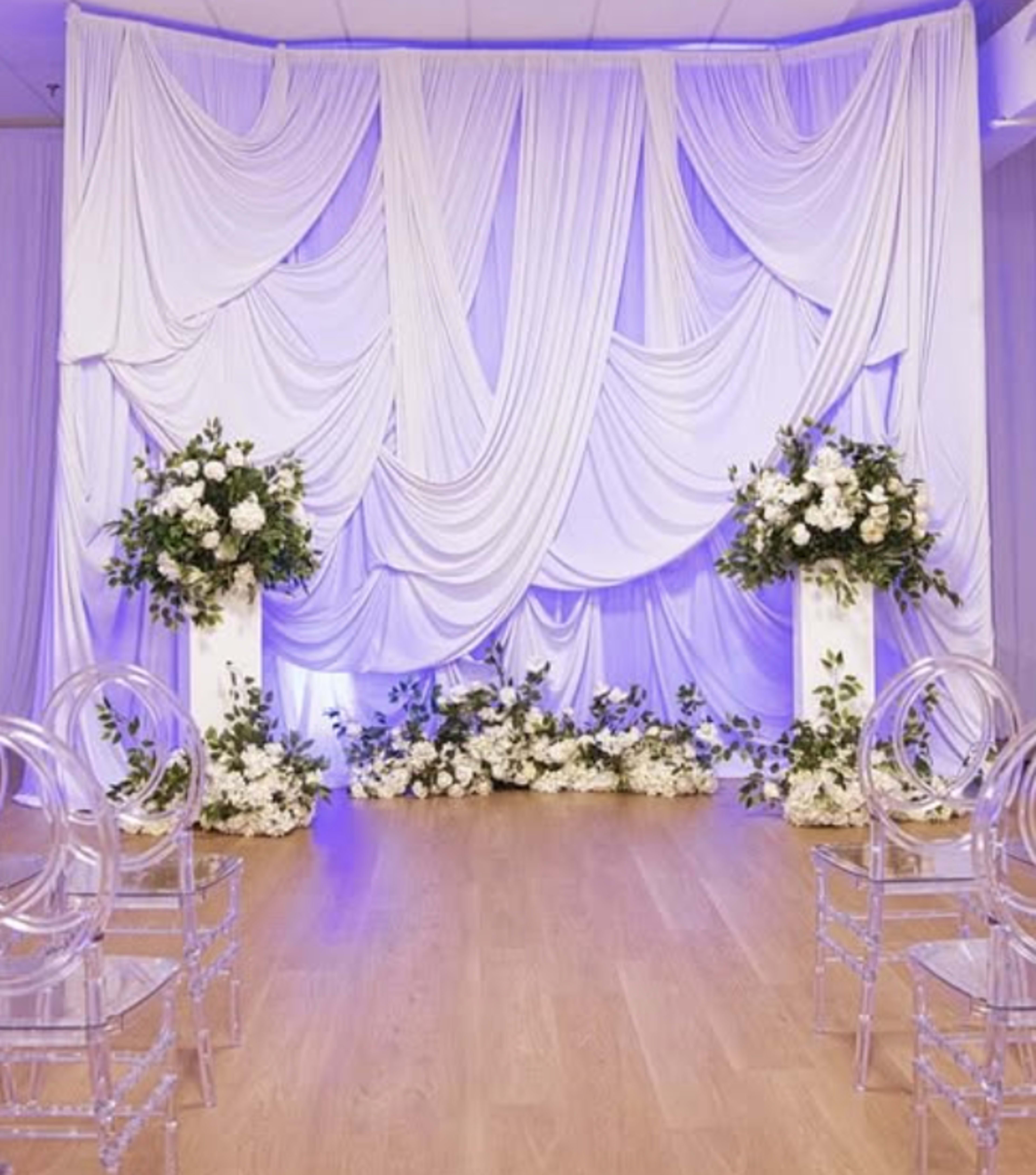The image features a wedding ceremony setup with a draped white fabric backdrop and floral arrangements on two pedestals, flanked by transparent chairs on a wooden floor.