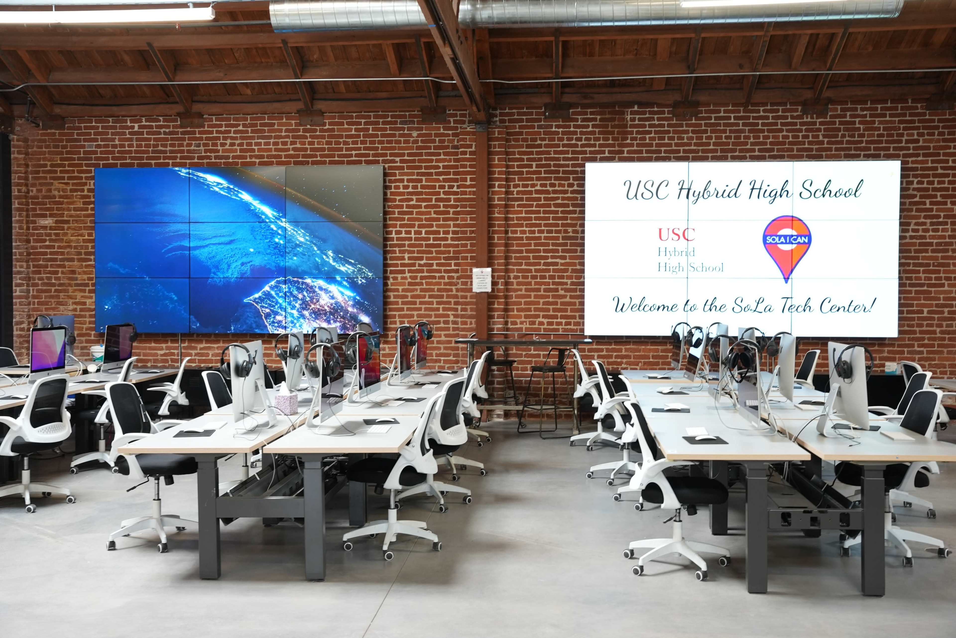 A modern classroom at USC Hybrid High School, featuring rows of desks equipped with computers and large screens displaying a world map and welcome text.