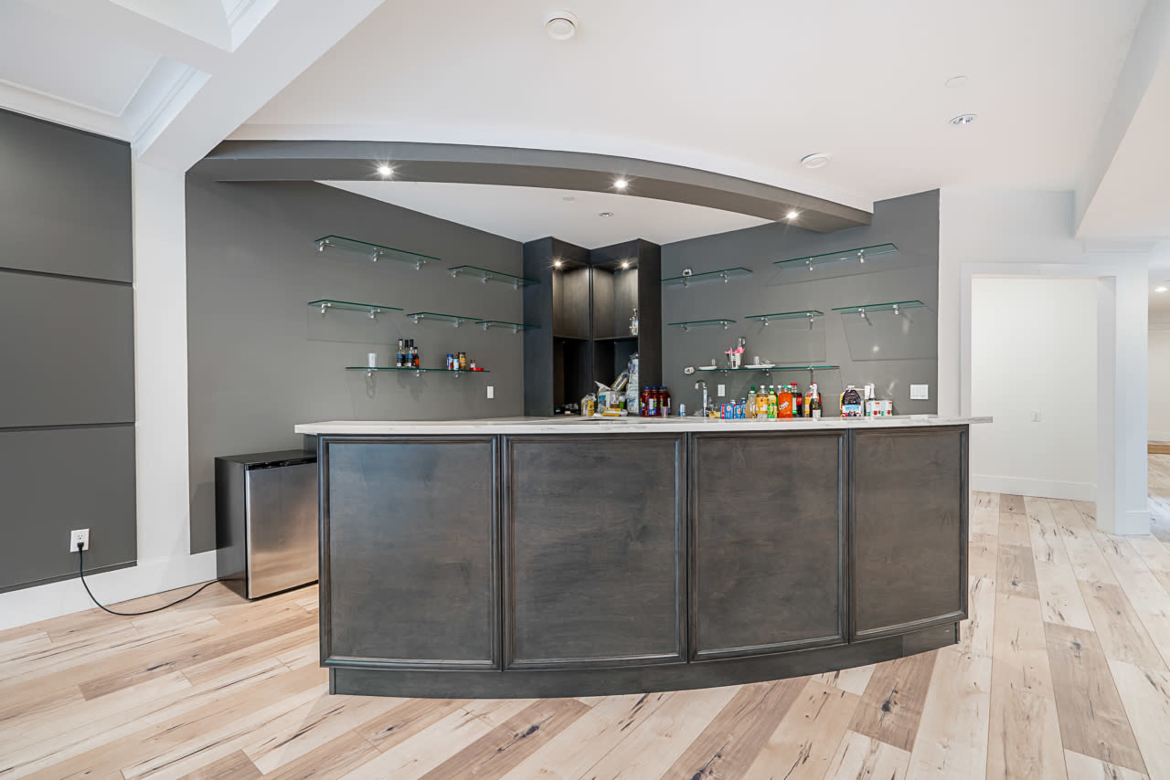 A modern home bar with a curved wooden countertop, mounted shelves displaying various bottles, and a minimalist design in a room with light wood flooring.