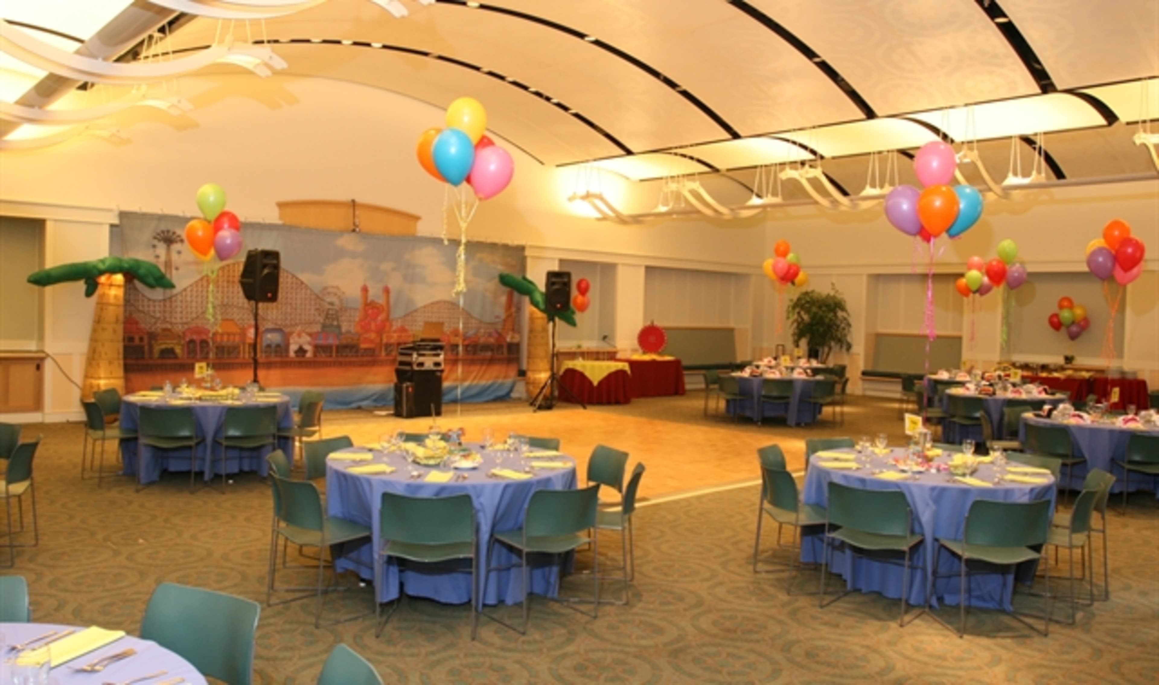 A spacious banquet hall features round tables with blue tablecloths, colorful balloons, and a staged backdrop with a cityscape.