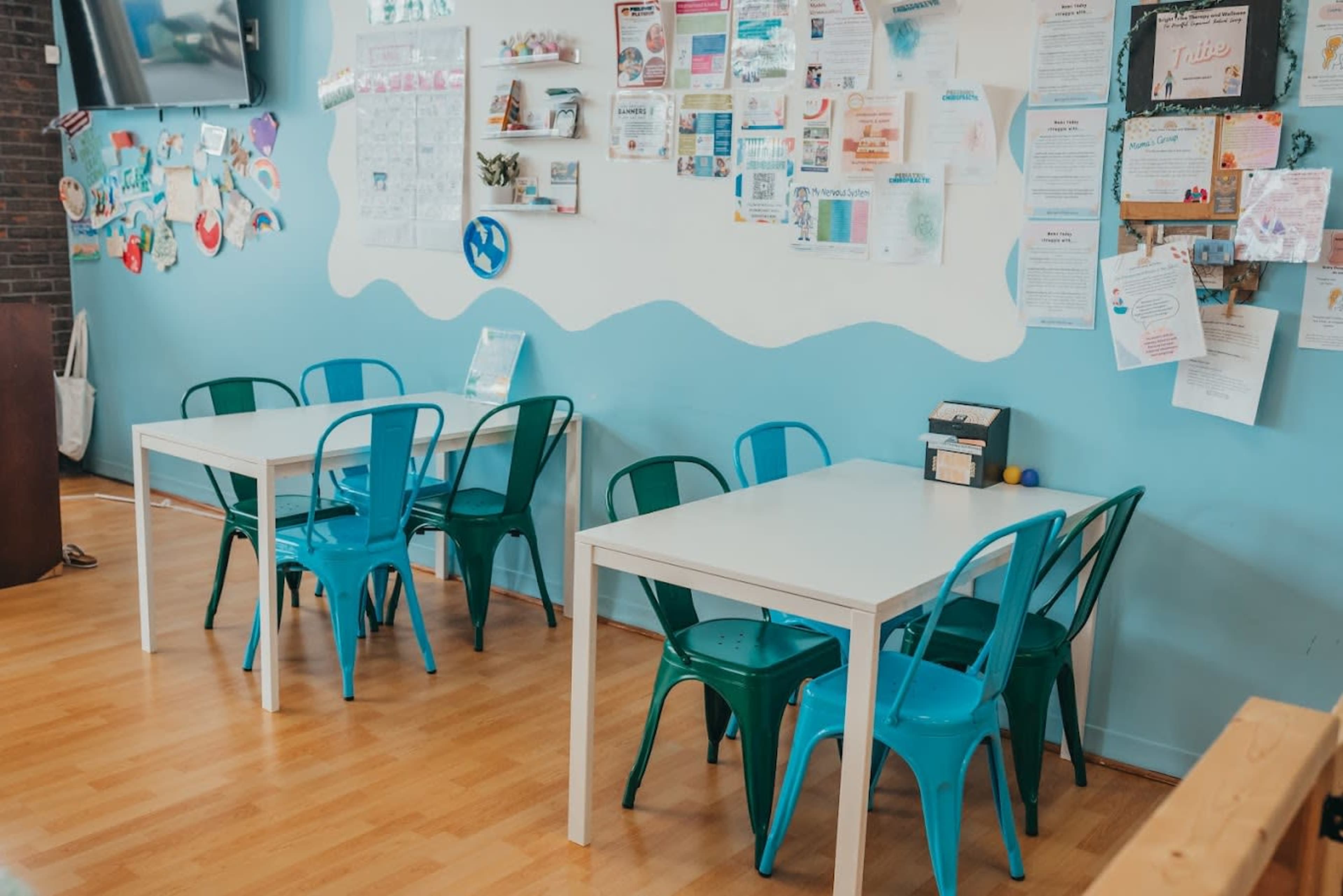 The image shows a bright indoor space with several tables and chairs against a blue wall adorned with various papers and notices.