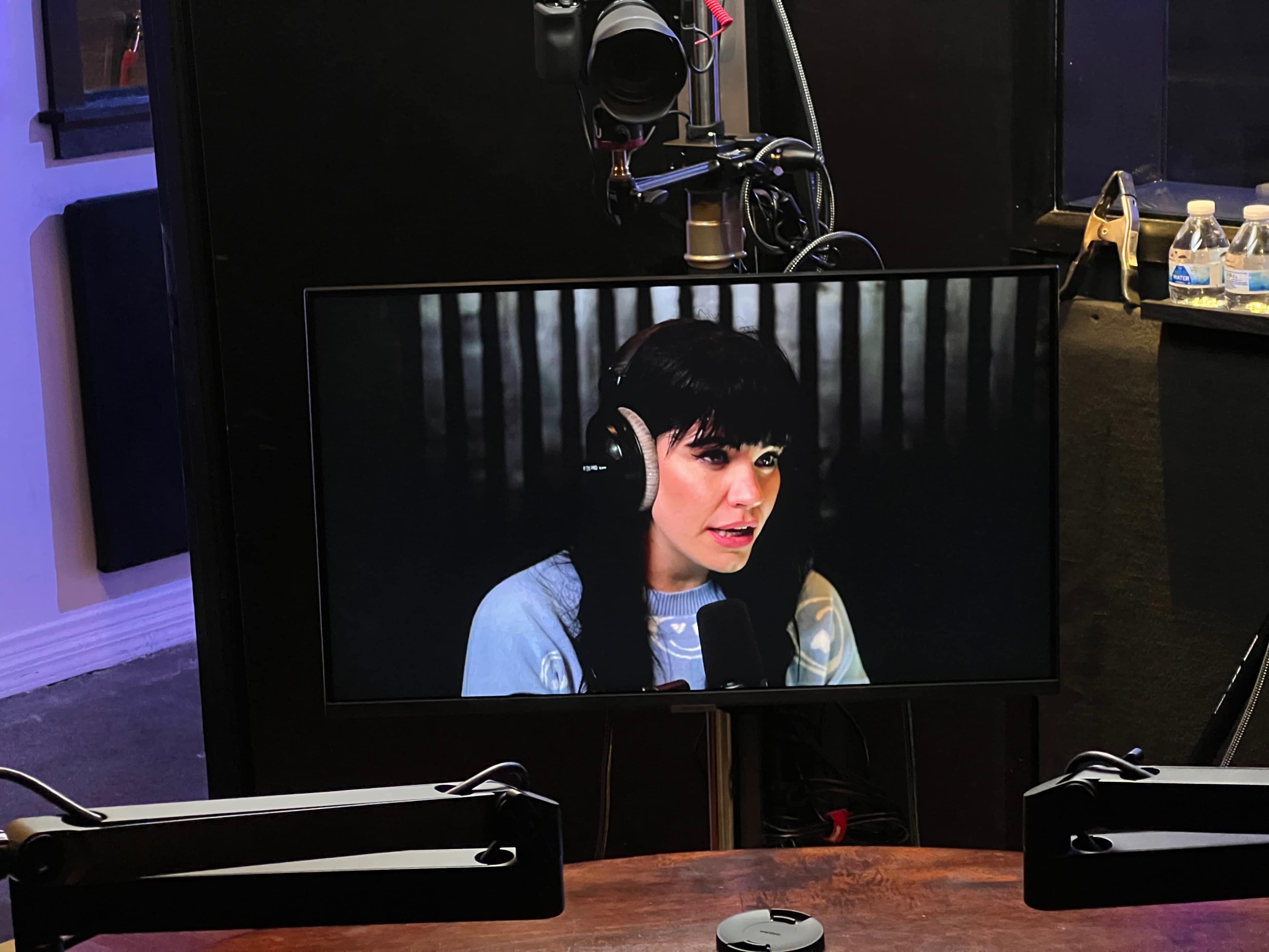 A woman with dark hair is speaking into a microphone while being filmed, displayed on a monitor in a recording studio.