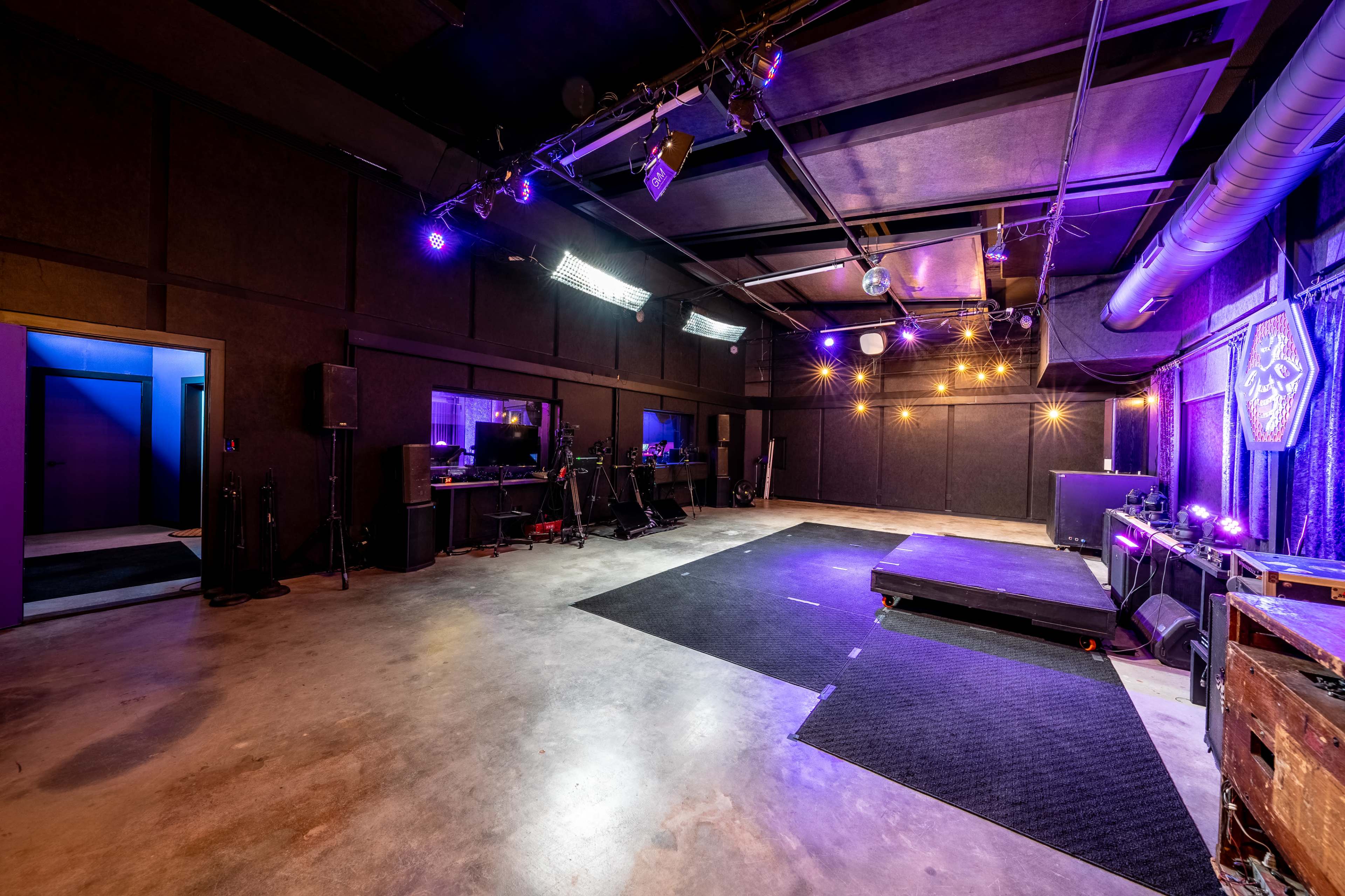 The image shows a spacious, dimly lit performance studio with black walls, stage lighting, and a small elevated stage in the center.