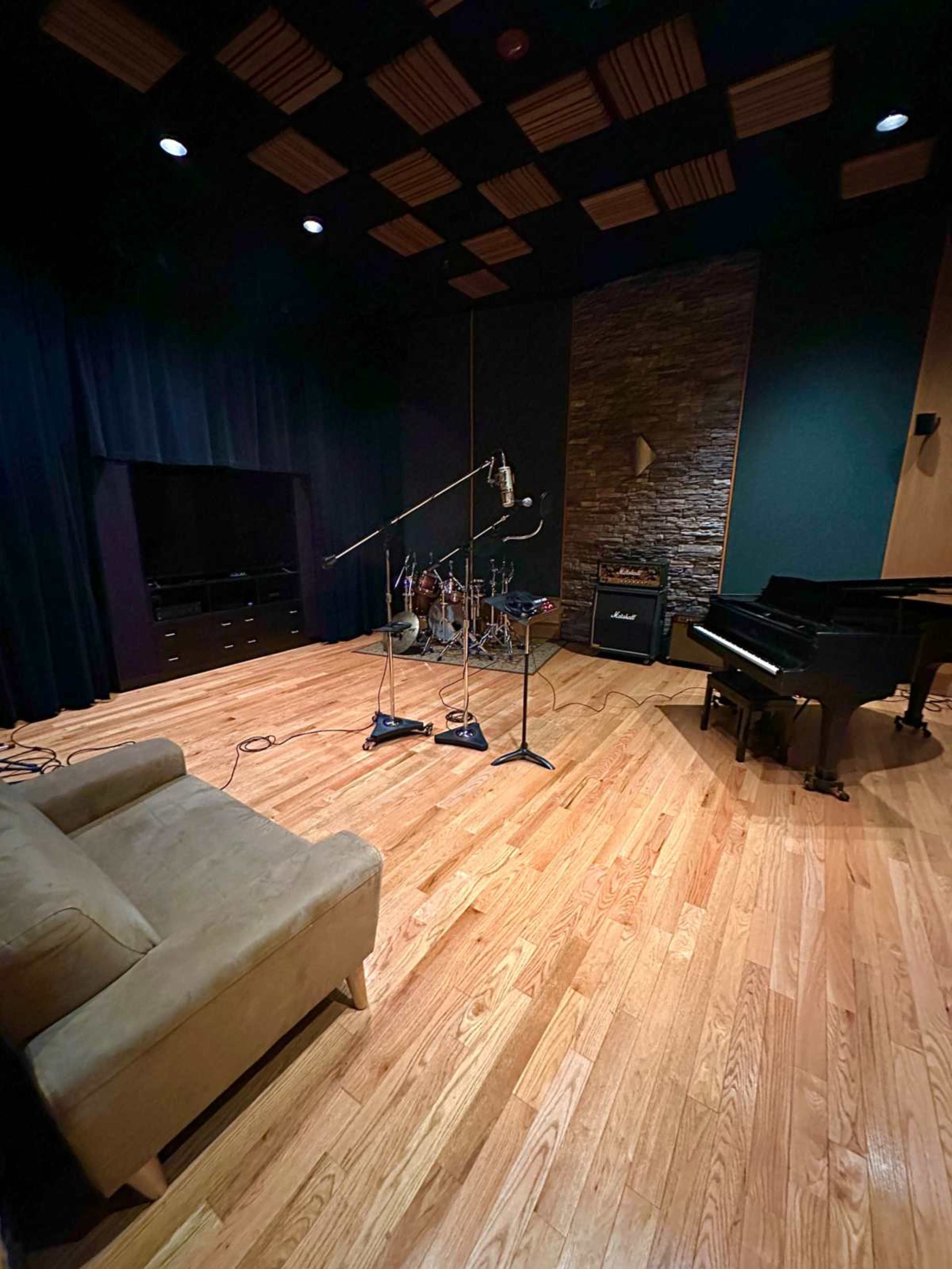 A music studio with a drum set, a grand piano, and microphones set up on a wooden floor.