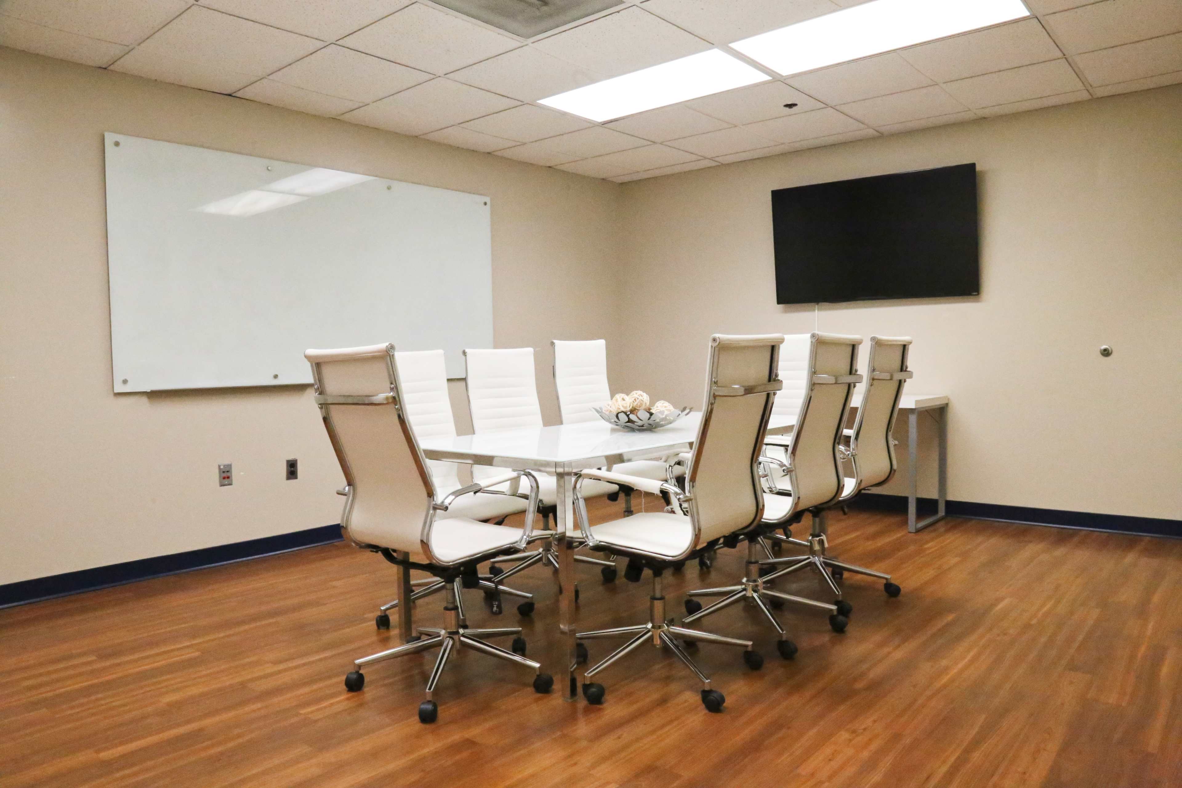 A modern conference room features a large glass table surrounded by eight white swivel chairs, with a wall-mounted TV and a whiteboard visible in the background.
