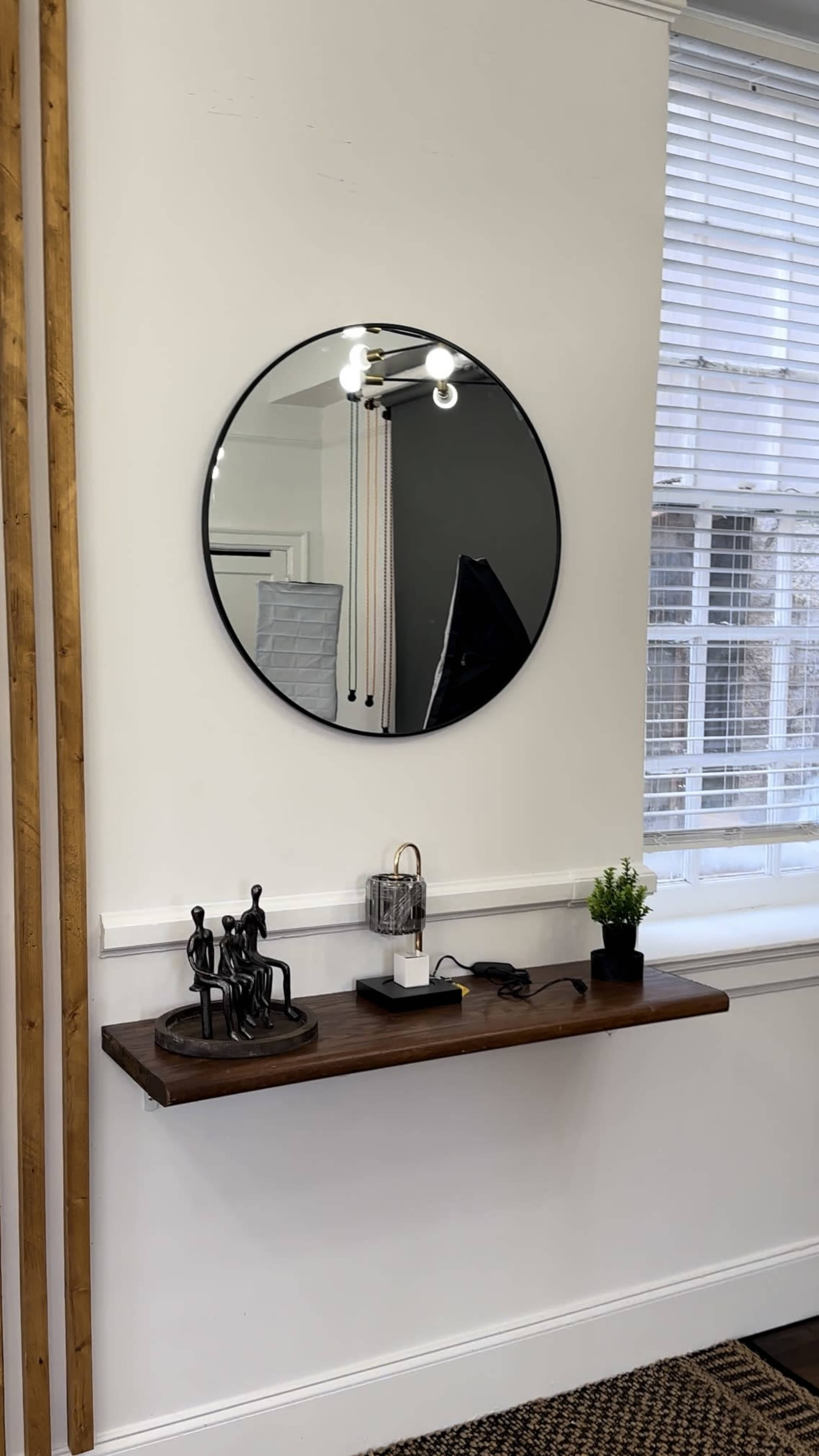 A round mirror hangs above a wooden shelf with decorative figures, a lamp, and a small plant against a white wall with natural light coming through a nearby window.