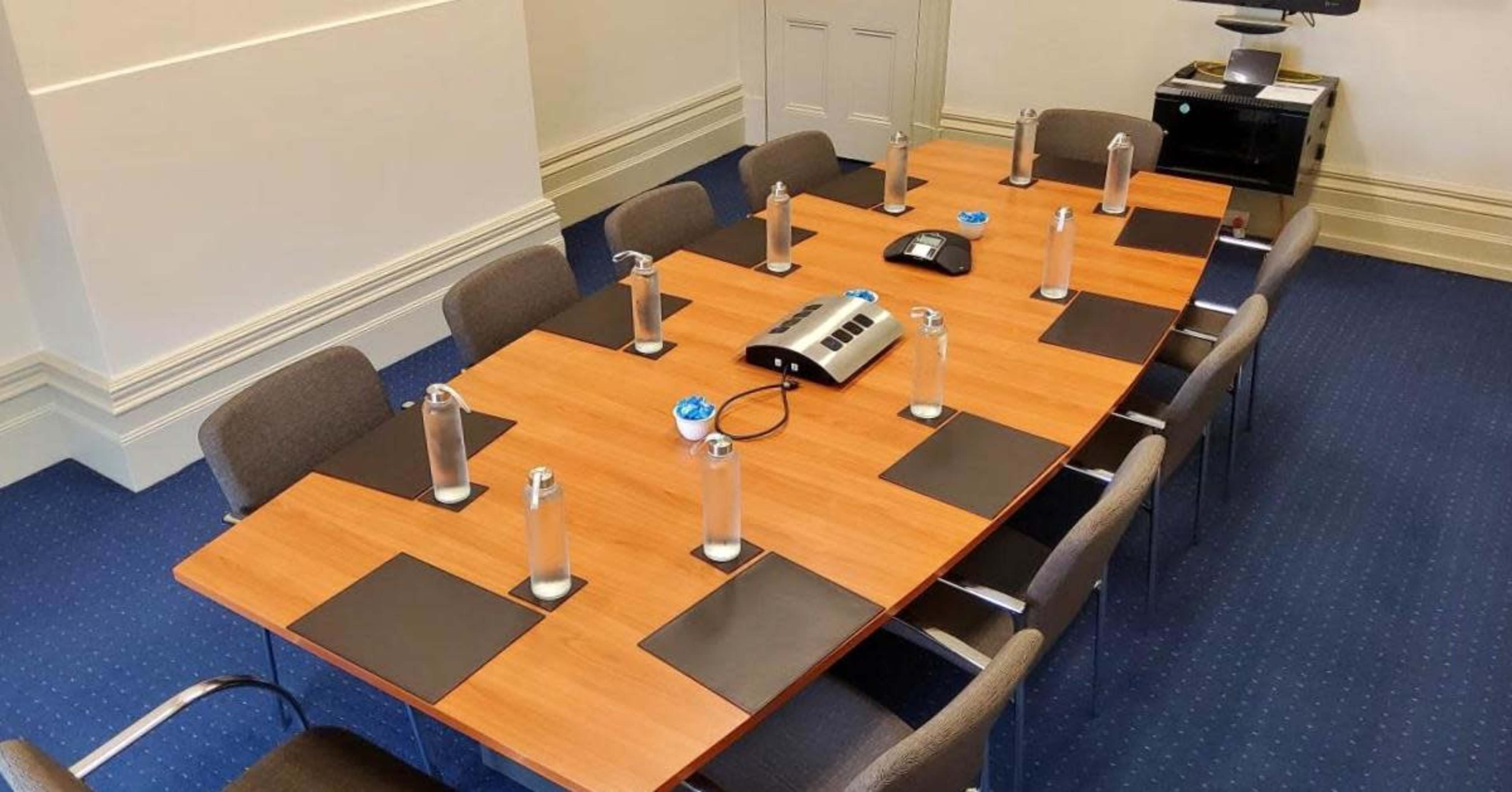A long wooden conference table is set up with ten chairs, each equipped with a water bottle and a notepad, and a speakerphone is positioned in the center.
