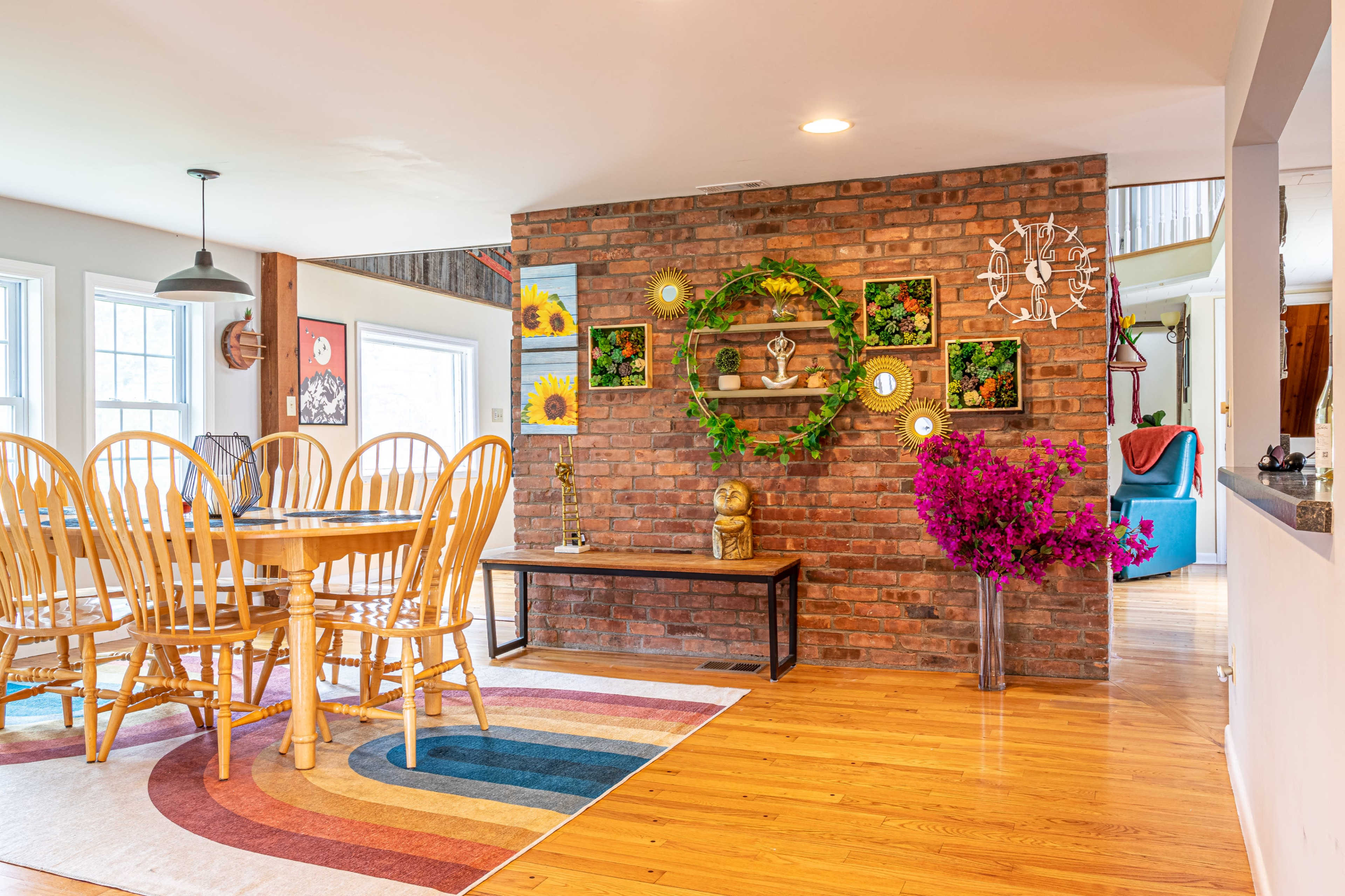 A dining area featuring a circular wooden table surrounded by six wooden chairs, a brick accent wall adorned with decorative items, and a colorful area rug.