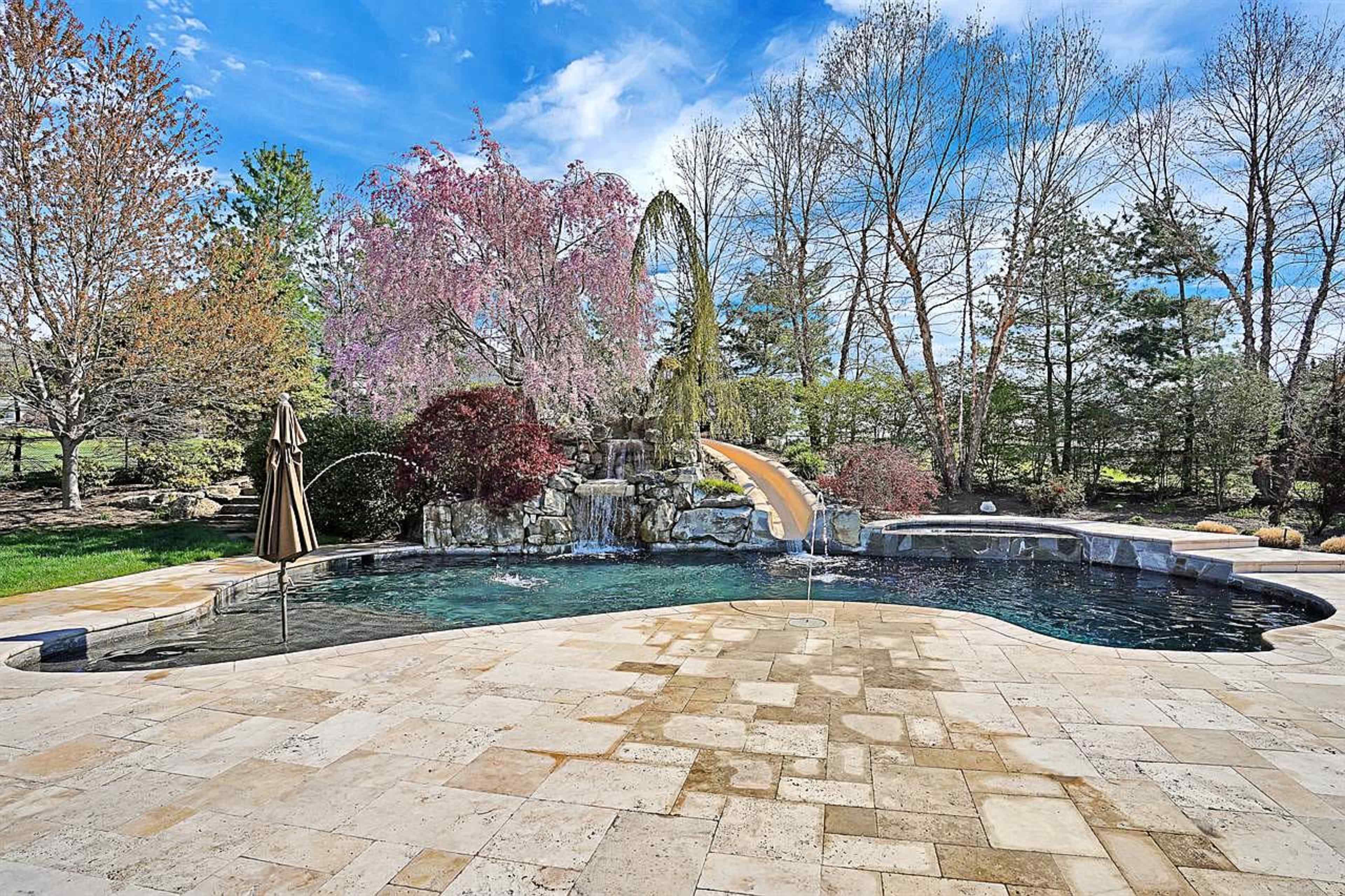 A landscaped backyard featuring a swimming pool with a waterfall and several trees, including a cherry blossom tree.