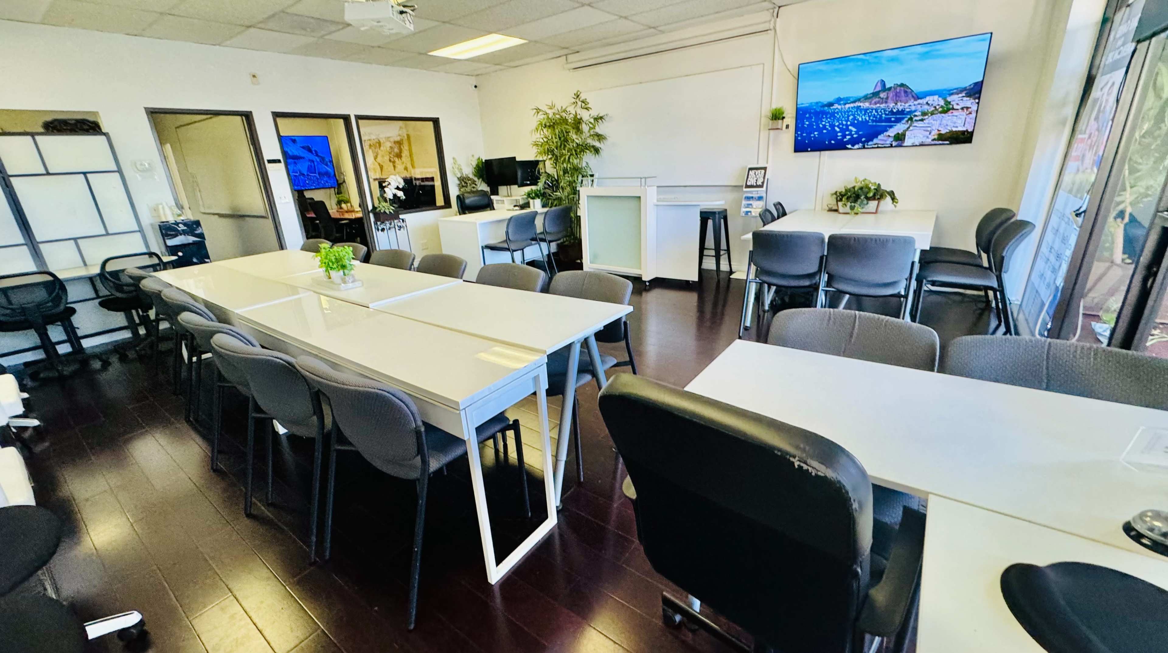 The image shows a modern office space with several white tables and gray chairs, large windows, and multiple screens displaying images on the walls.